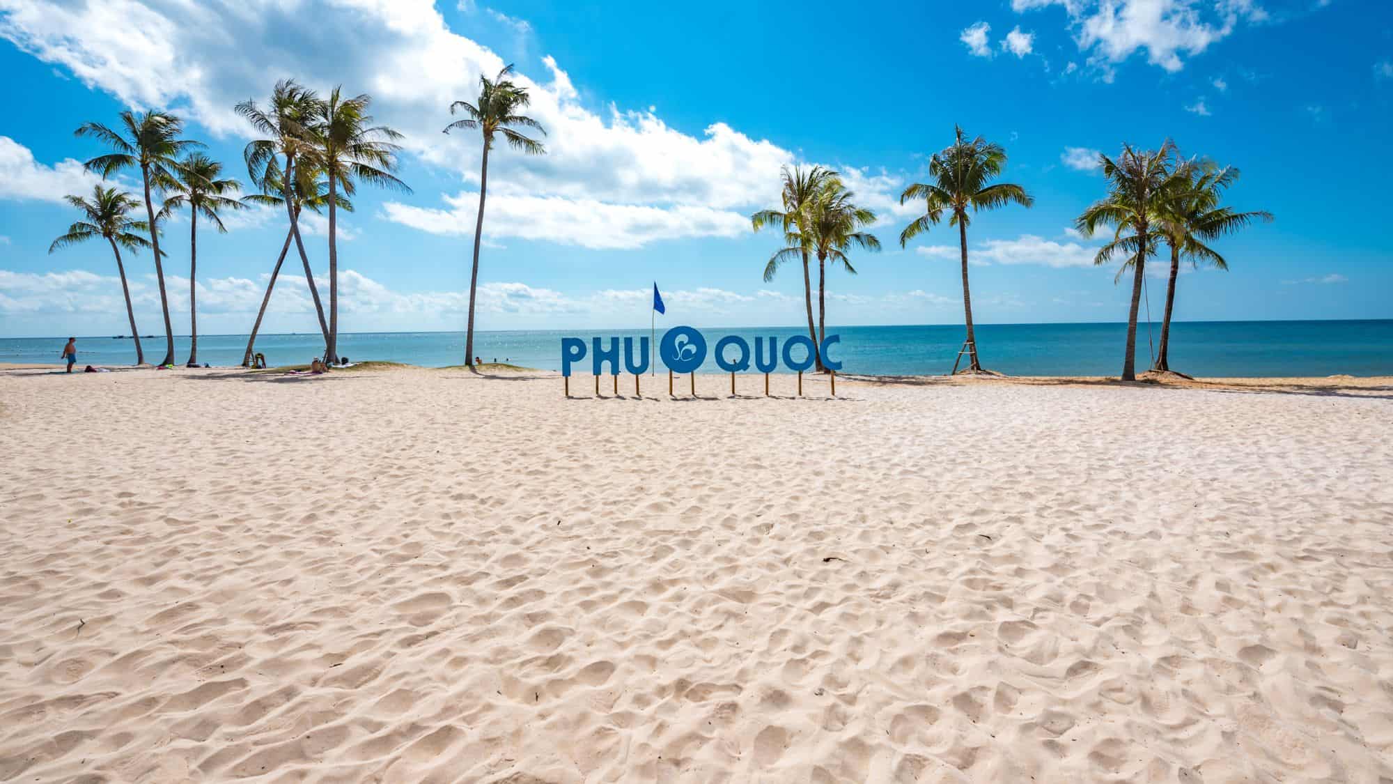 A pristine sandy beach with tall palm trees and a large “Phu Quoc” sign standing against the deep blue ocean and a bright sky.