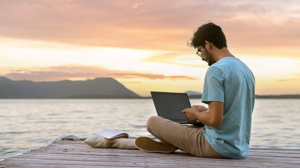 A young man in a casual blue t-shirt and glasses sits cross-legged on a wooden dock, working on his laptop as the sun sets over a calm lake. A notebook and cushion rest beside him, enhancing the serene remote work setting.