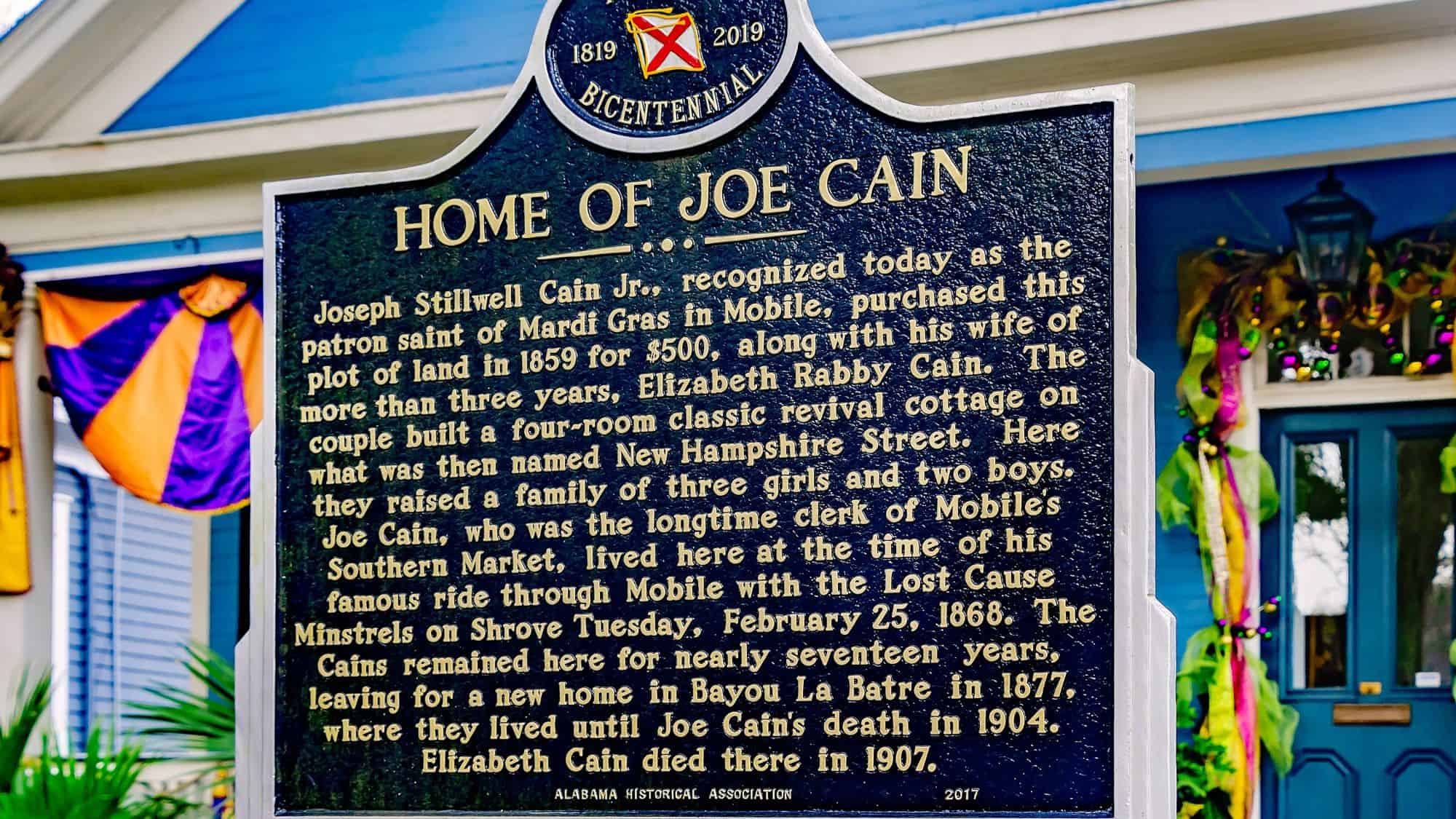 A historical plaque in front of a house in Mobile, Alabama, detailing the history of Joe Cain, the "patron saint of Mardi Gras in Mobile." The sign is surrounded by festive decorations in purple, green, and gold.