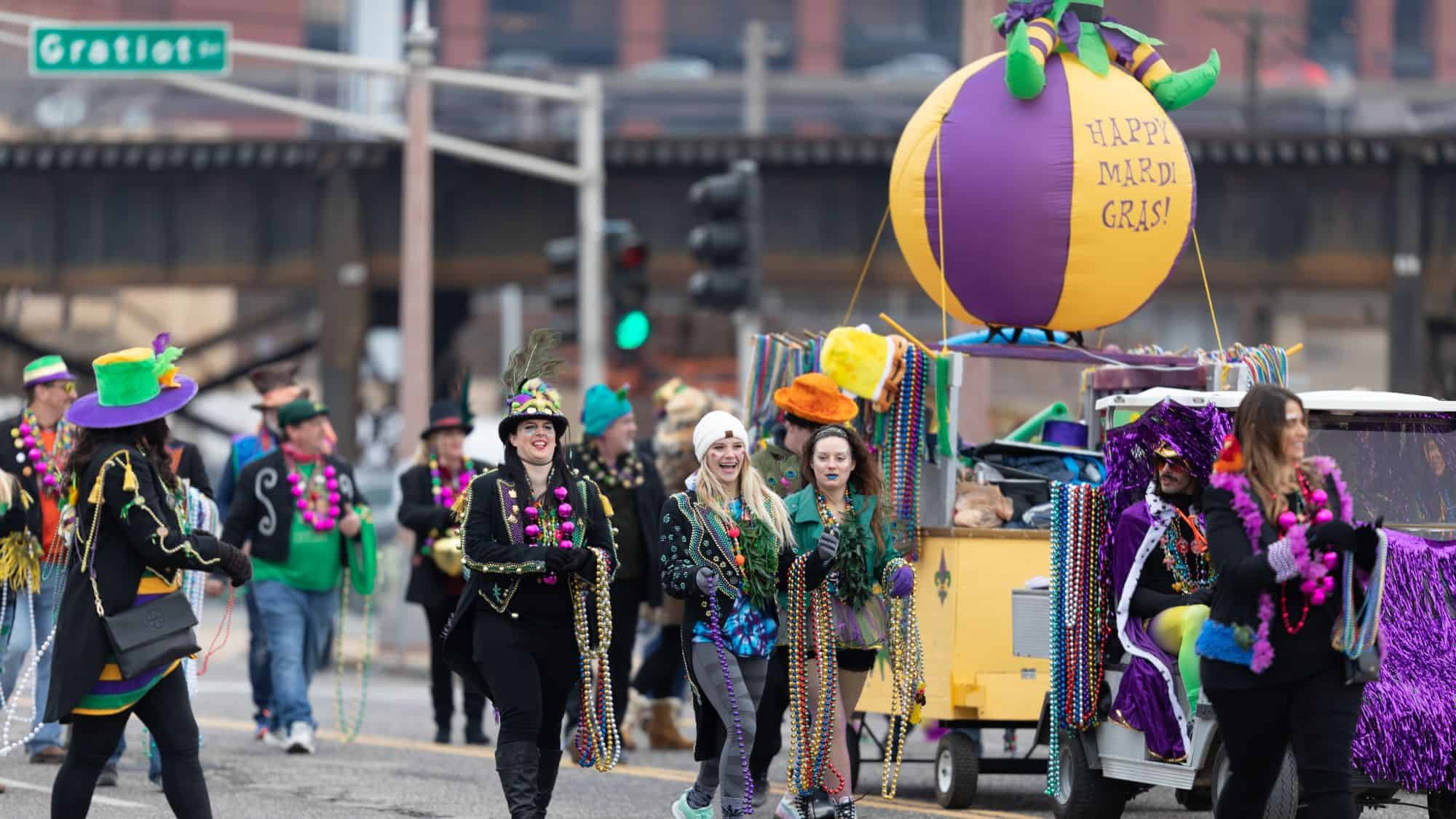 A lively street parade with participants dressed in colorful Mardi Gras outfits, wearing bead necklaces and feathered hats. A decorated float features a large inflatable ball with "Happy Mardi Gras!" written on it.