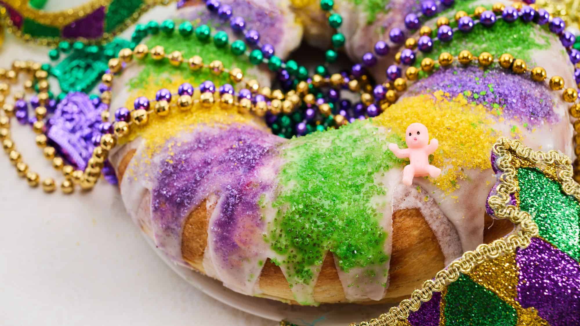 Close-up of a traditional Mardi Gras King Cake topped with white icing, purple, green, and gold sprinkles, adorned with shiny beads in matching colors, and featuring a small pink plastic baby on top.