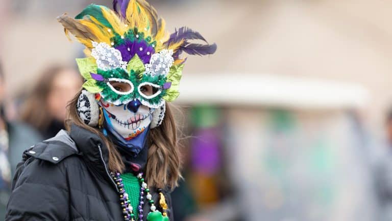 A person in a vibrant Mardi Gras mask decorated with feathers, sequins, and lace in green, purple, and gold. Their face is painted with a sugar skull design, and they are wearing festive beads around their neck.