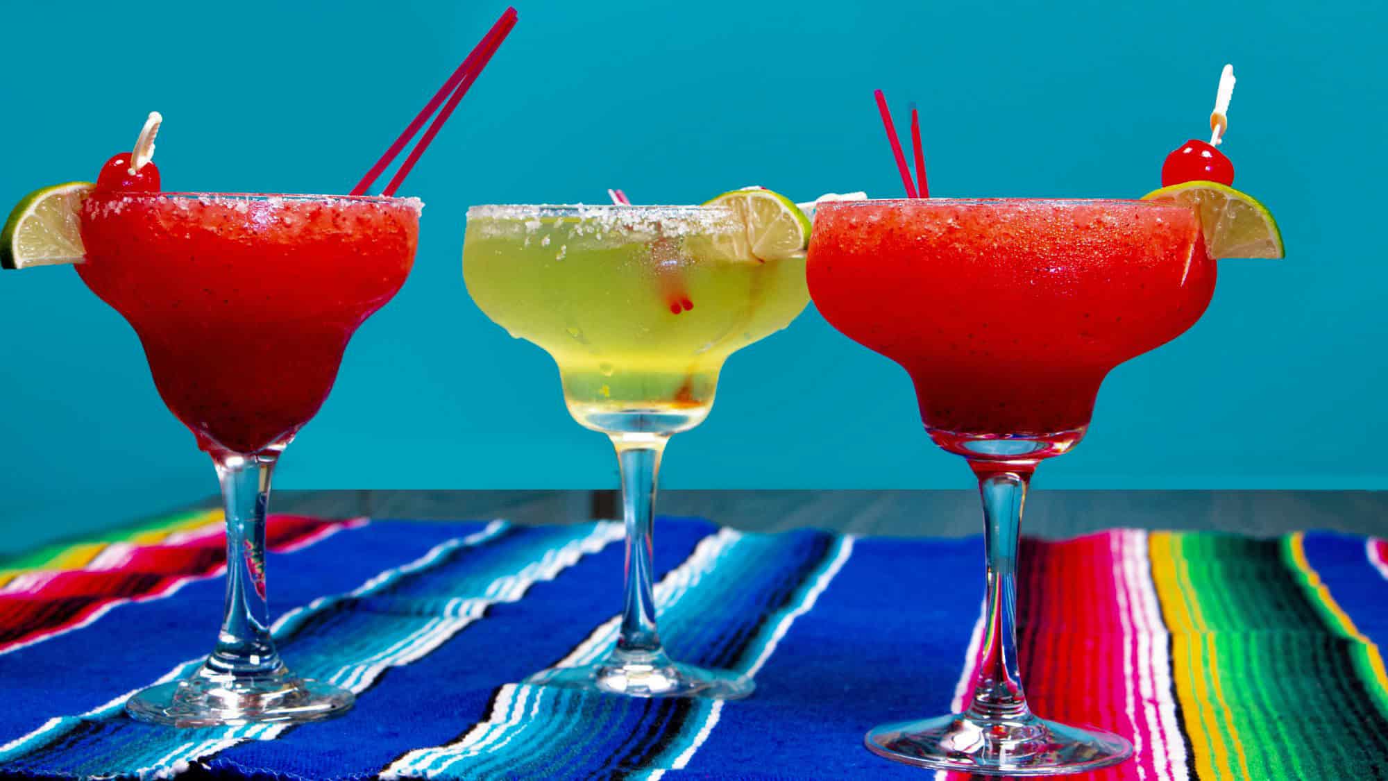 Three margarita glasses filled with bright red and green frozen drinks are displayed on a colorful striped Mexican blanket, garnished with lime wedges and cherries.