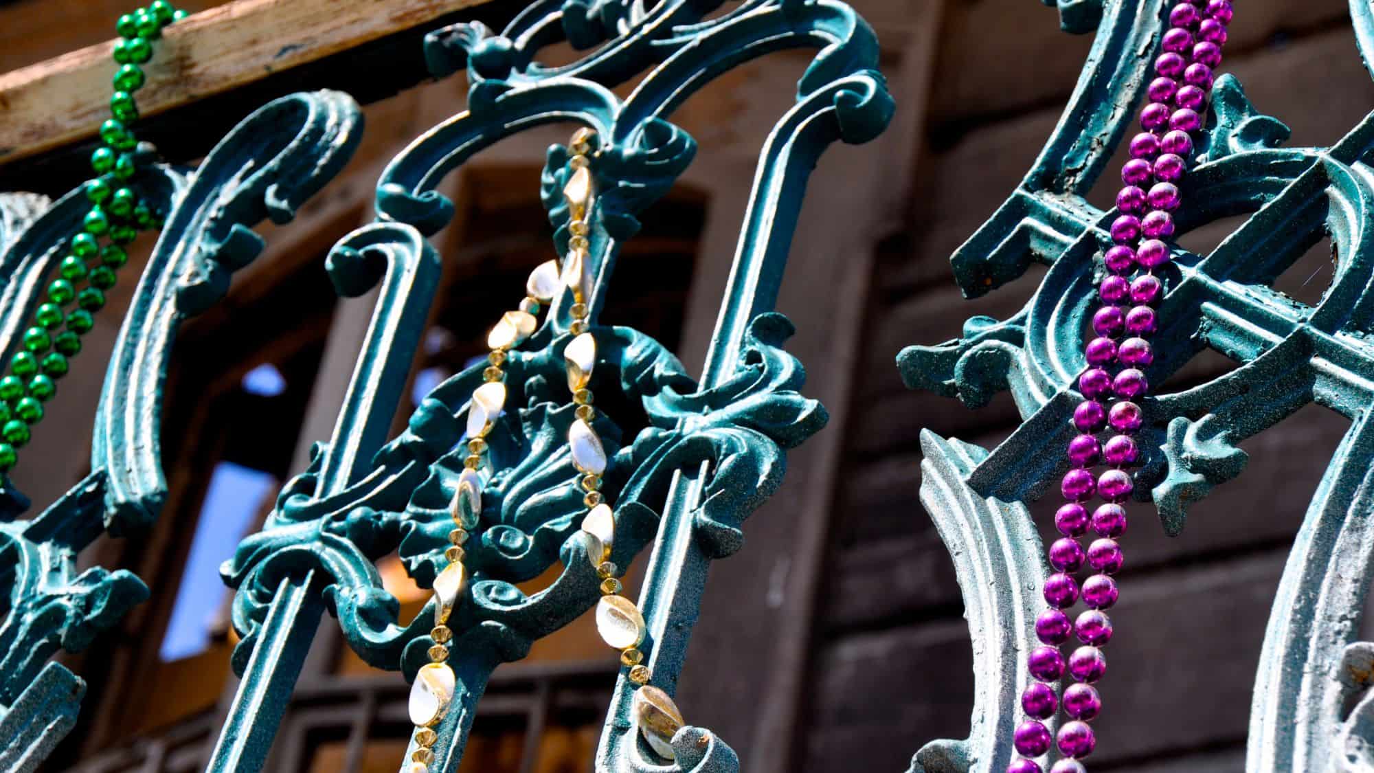 A close-up shot of an ornate wrought-iron fence adorned with colorful Mardi Gras beads in green, gold, and purple. The beads drape over the intricate metalwork, with blurred buildings in the background.