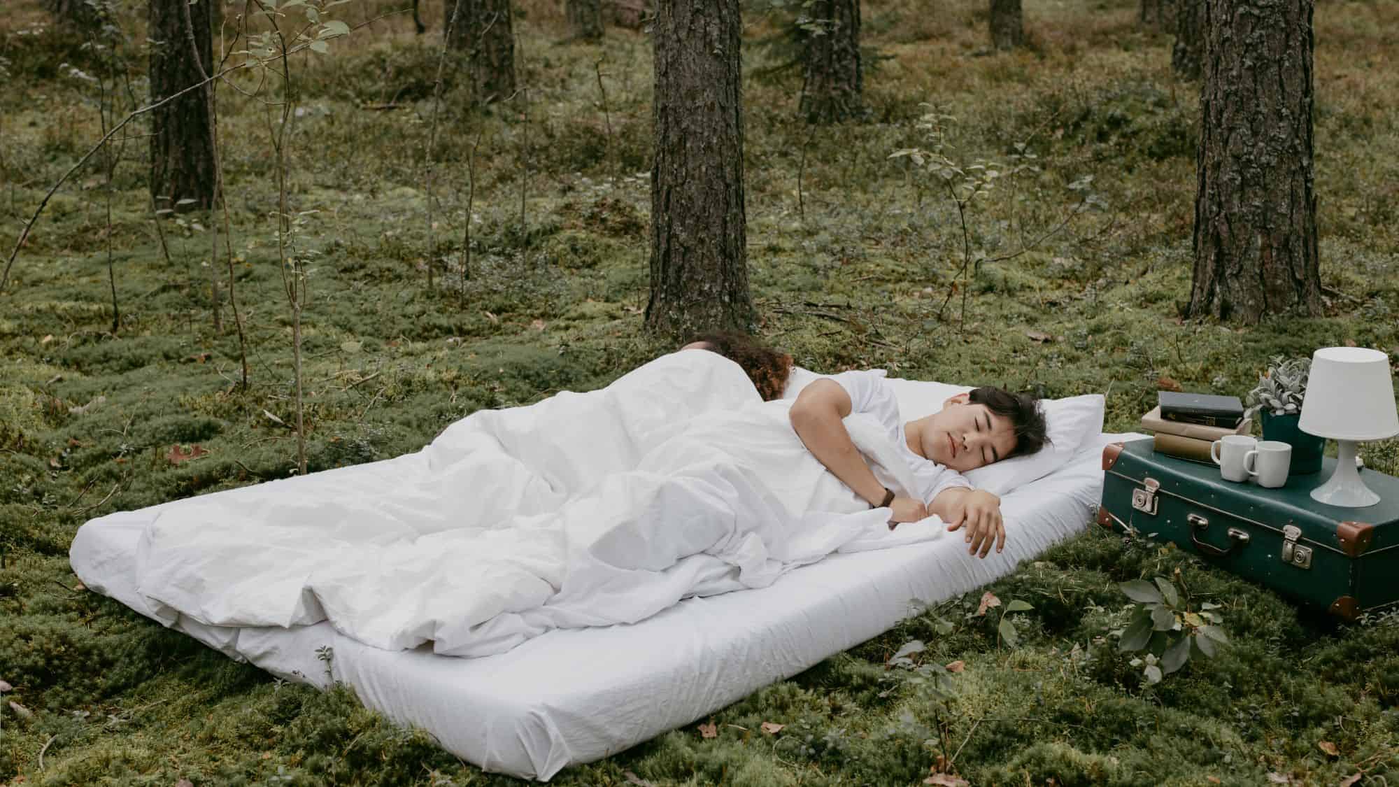 Two people sleep peacefully on a white mattress placed directly on a mossy forest floor. A small bedside setup with books, cups, and a lamp adds an unexpected homely touch.