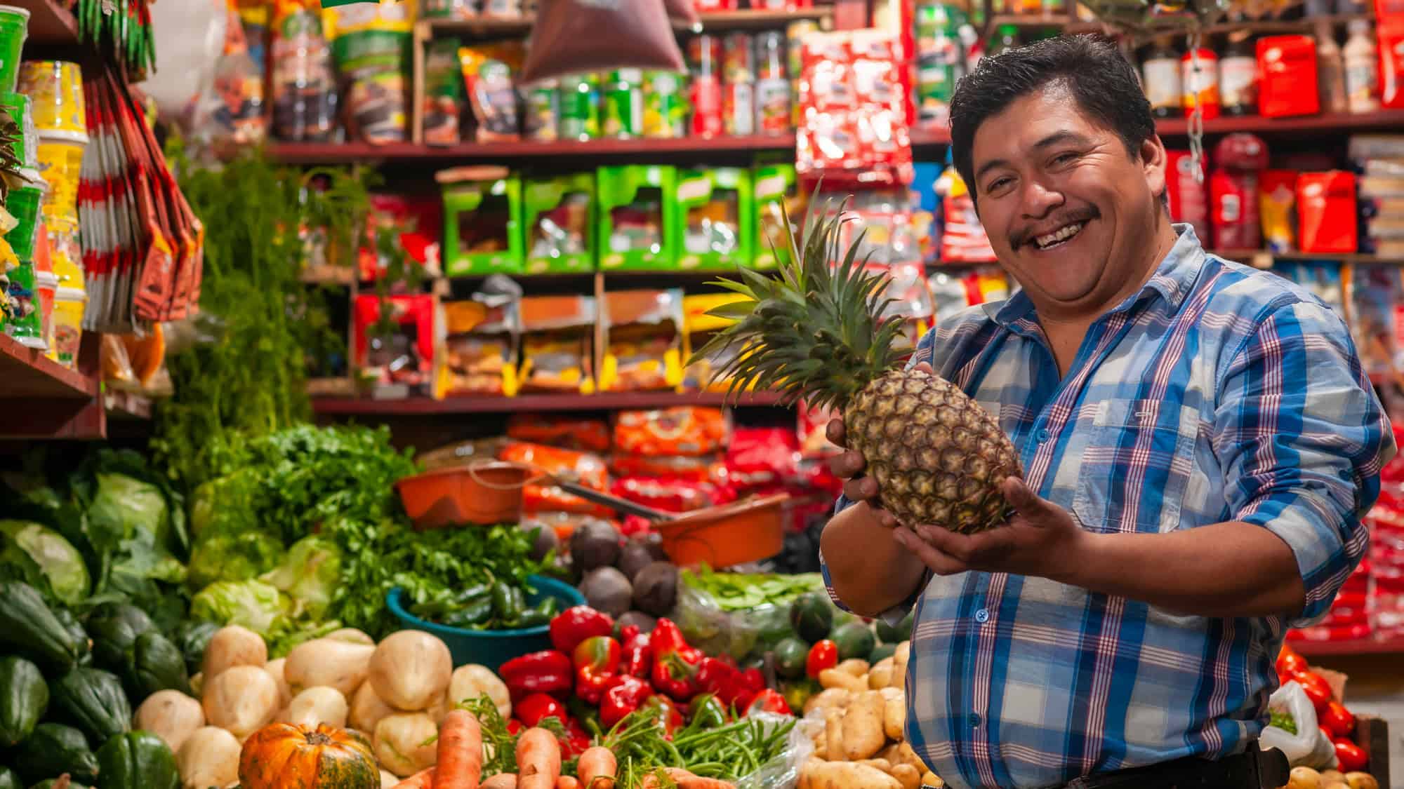 A cheerful vendor in a blue plaid shirt holds a pineapple while standing behind a colorful array of fresh fruits and vegetables in a busy market stall.