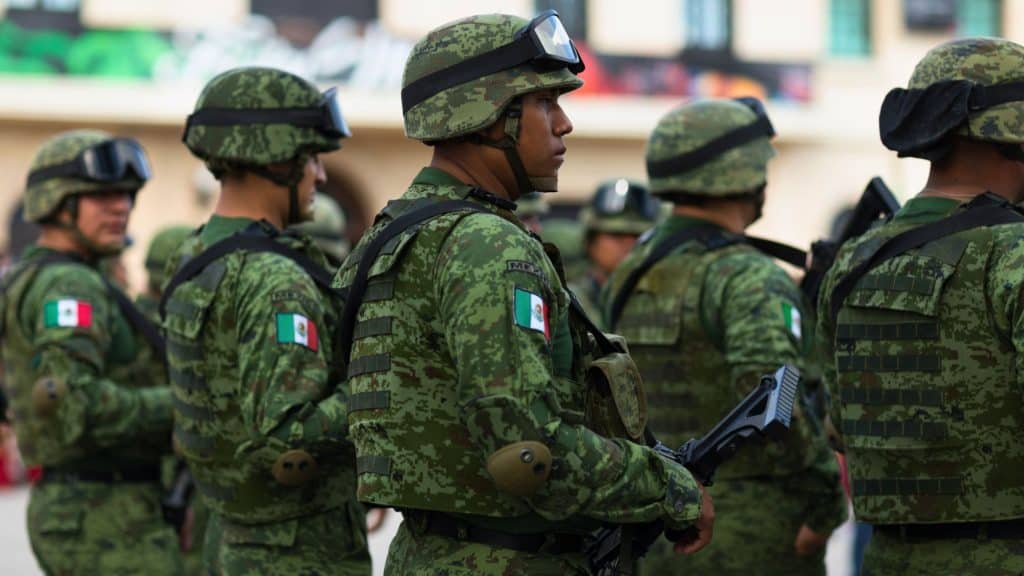 A group of Mexican soldiers in green camouflage uniforms and helmets standing in formation. The Mexican flag patch on their sleeves is visible as they carry rifles, looking focused and prepared.