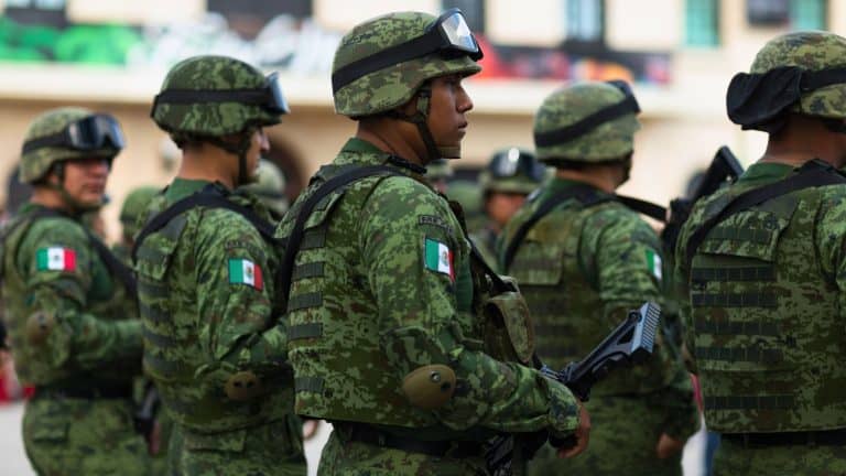 A group of Mexican soldiers in green camouflage uniforms and helmets standing in formation. The Mexican flag patch on their sleeves is visible as they carry rifles, looking focused and prepared.