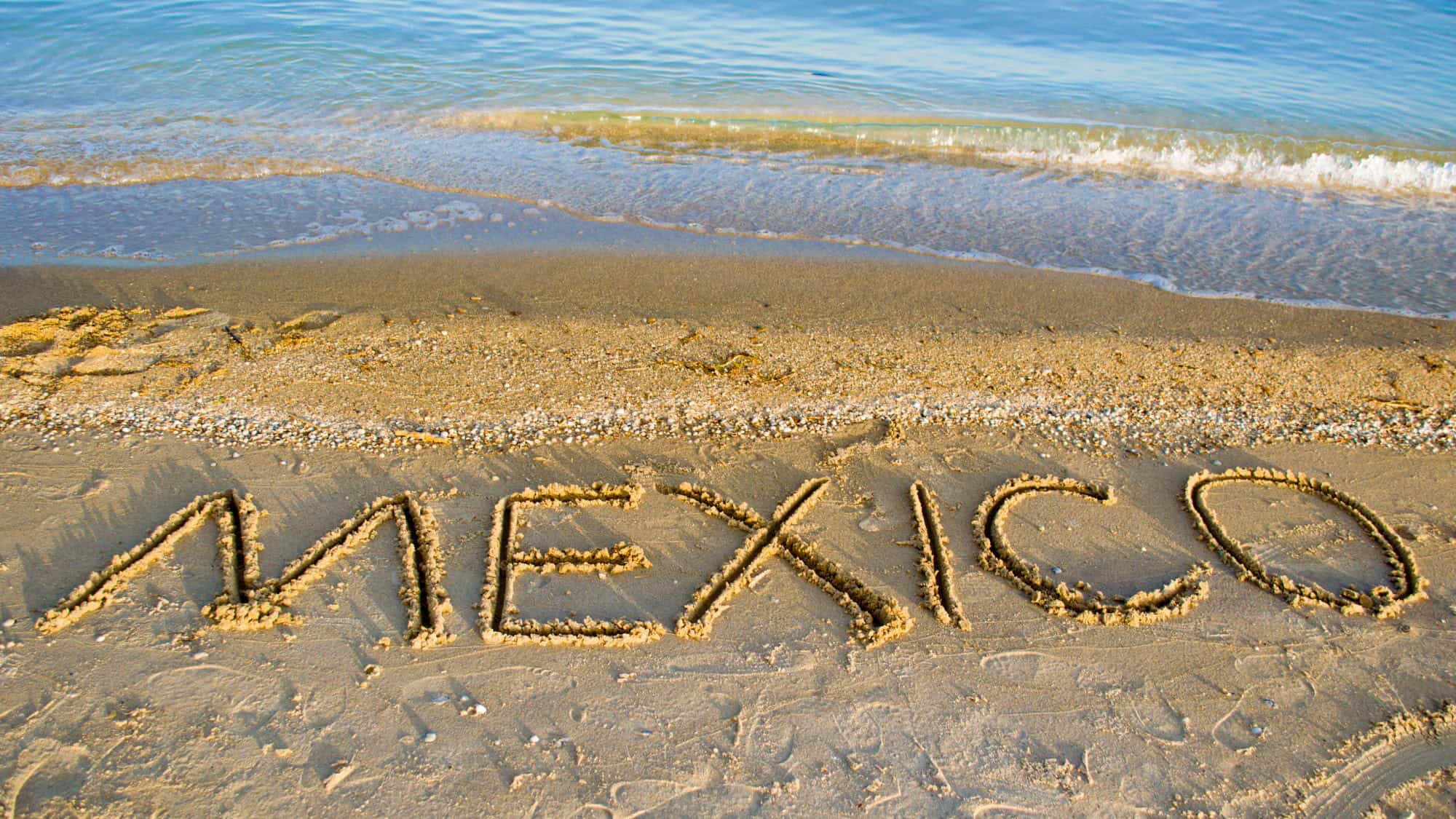 The word "MEXICO" is written in large, bold letters in the wet sand along a beach, with gentle waves lapping at the shore in the background.