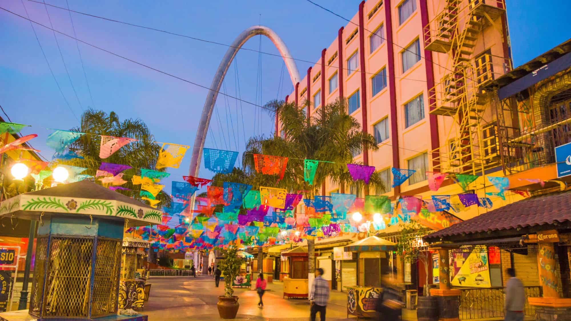 A vibrant street scene in Tijuana, Mexico, featuring colorful papel picado (decorative paper banners) hanging over a pedestrian street. The Millennial Arch, a large metallic structure, rises in the background, with pastel-colored buildings and palm trees adding to the lively atmosphere.