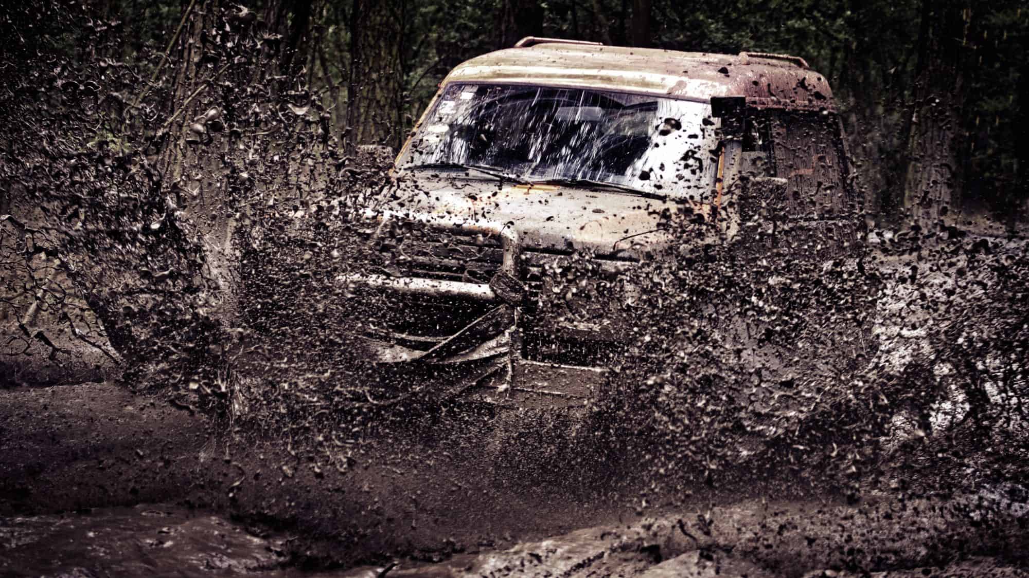 An off-road SUV plowing through thick mud, creating a dramatic spray of dirt and water. The vehicle is covered in mud, emphasizing the intensity of the rugged terrain.