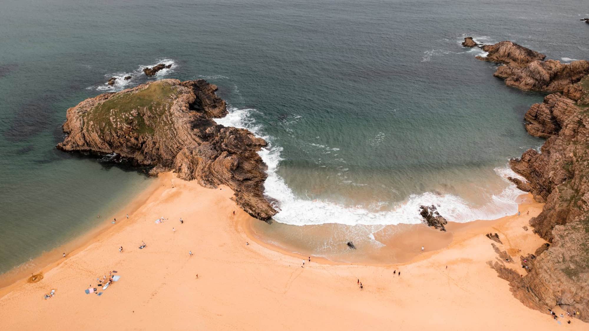 A secluded golden sand beach with jagged rock formations and turquoise waves crashing against the shore. A few visitors are scattered along the sand, adding a sense of adventure to the rugged coastline.