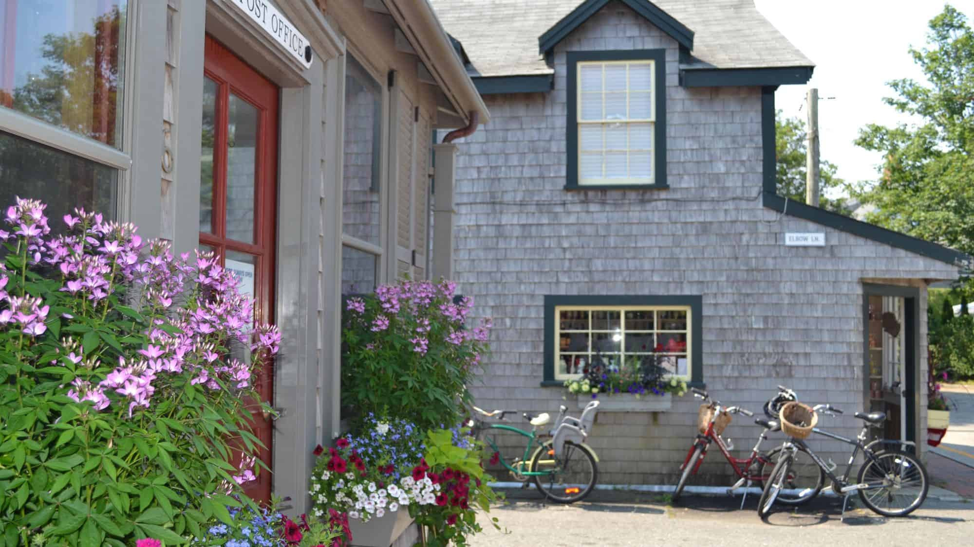 A charming street scene in Nantucket, Massachusetts, featuring weathered gray shingle buildings, blooming purple flowers, and several bicycles parked along the sidewalk, evoking a classic coastal New England vibe.