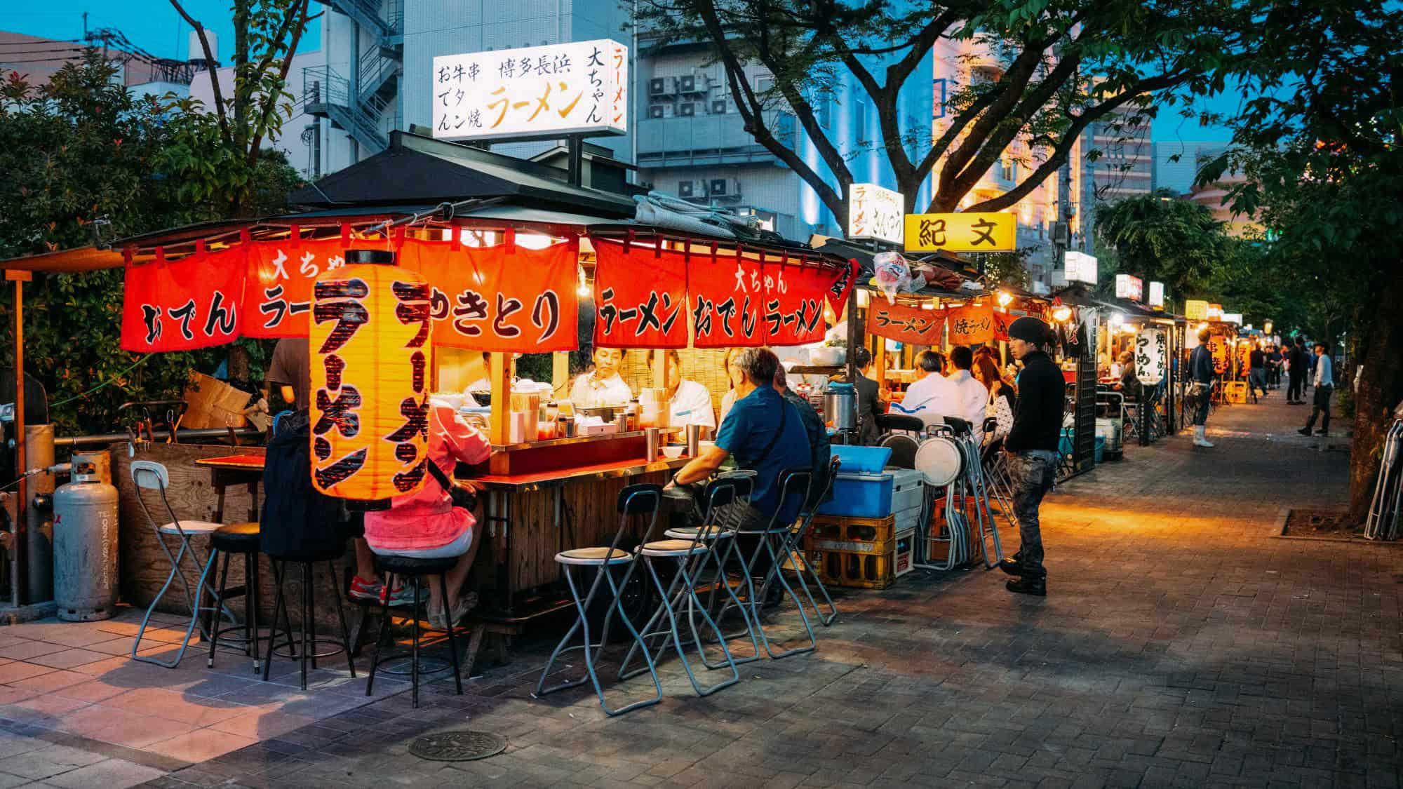A lively nighttime scene of an outdoor ramen street food market in Japan. People are sitting on stools, enjoying hot bowls of ramen under bright red lanterns, with chefs preparing meals inside the small stalls.