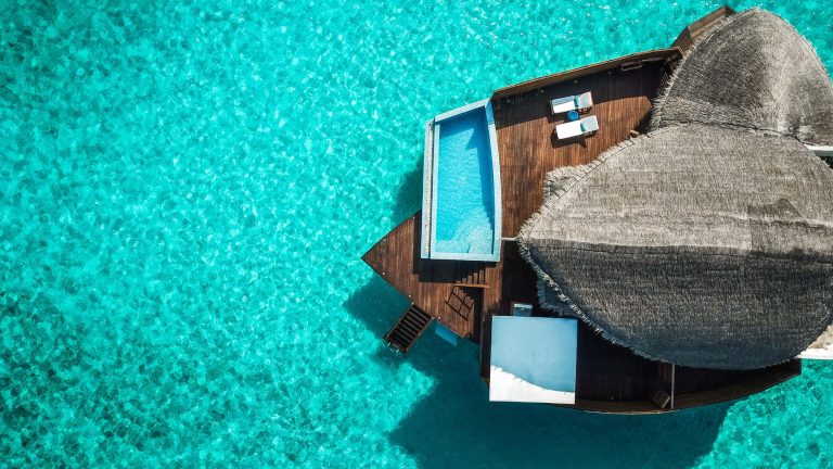 Close-up aerial shot of an overwater villa featuring a private pool, sun loungers, and a thatched roof, all surrounded by crystal-clear waters. A small staircase leads directly into the ocean.