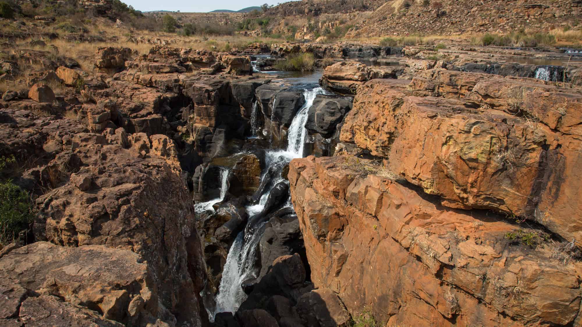A rugged landscape featuring cascading waterfalls flowing through rocky formations. The reddish-brown rock contrasts with the white, rushing water, making for a striking natural scene.