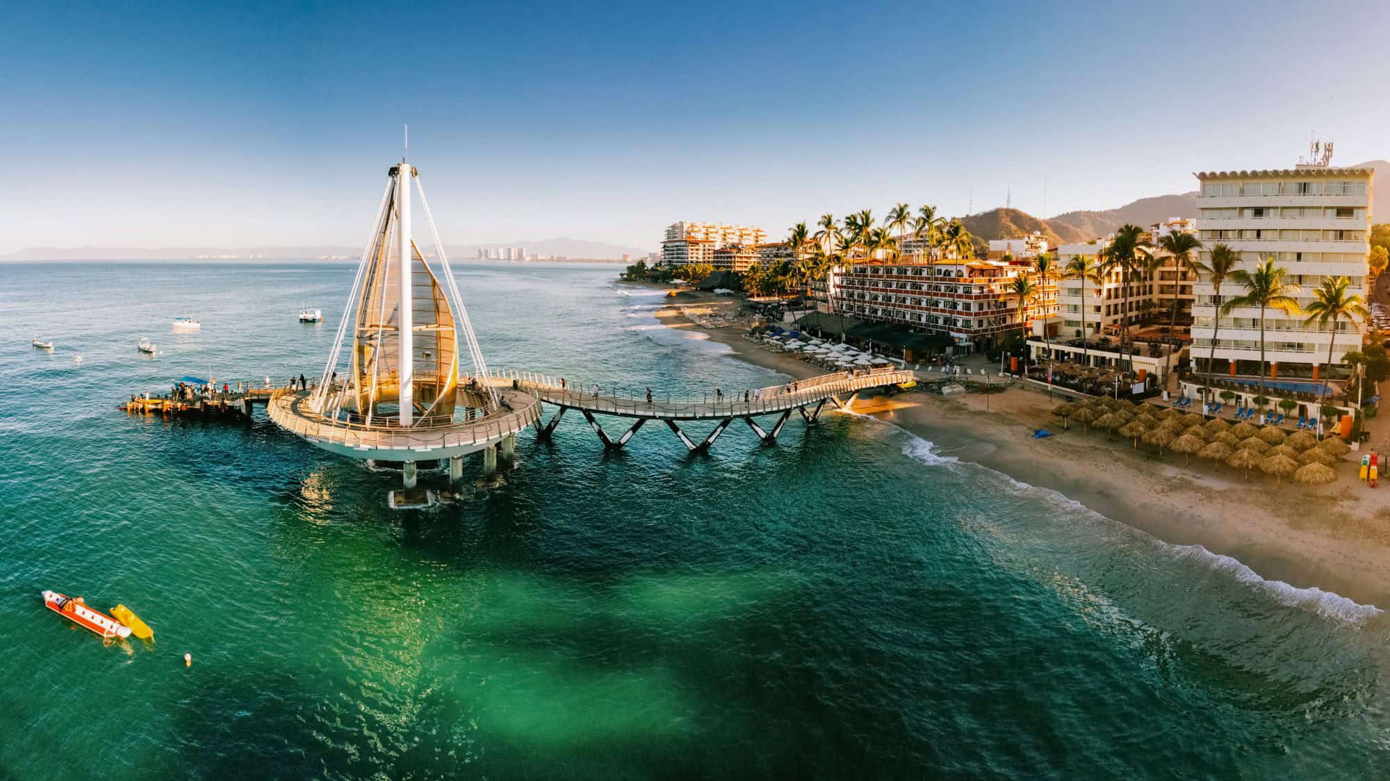 The Los Muertos Pier in Puerto Vallarta extends over turquoise waters, surrounded by small boats and beachfront hotels under a clear sky.