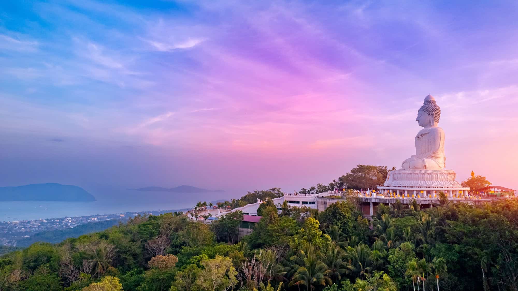 The Big Buddha of Phuket sits atop lush greenery, overlooking the island at sunset with a sky painted in pastel hues of pink, purple, and blue.