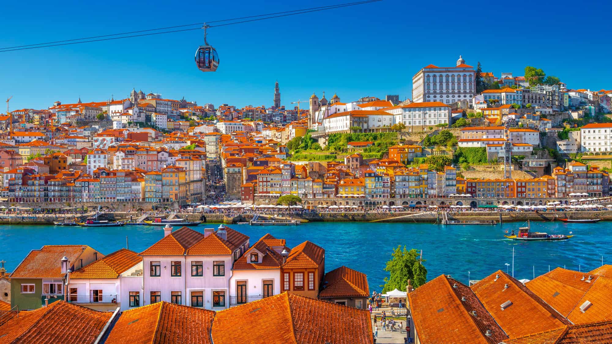 A colorful panoramic view of Porto, Portugal, with its famous terracotta-roofed buildings lining the banks of the Douro River. A cable car glides above the city, adding to the picturesque European charm.