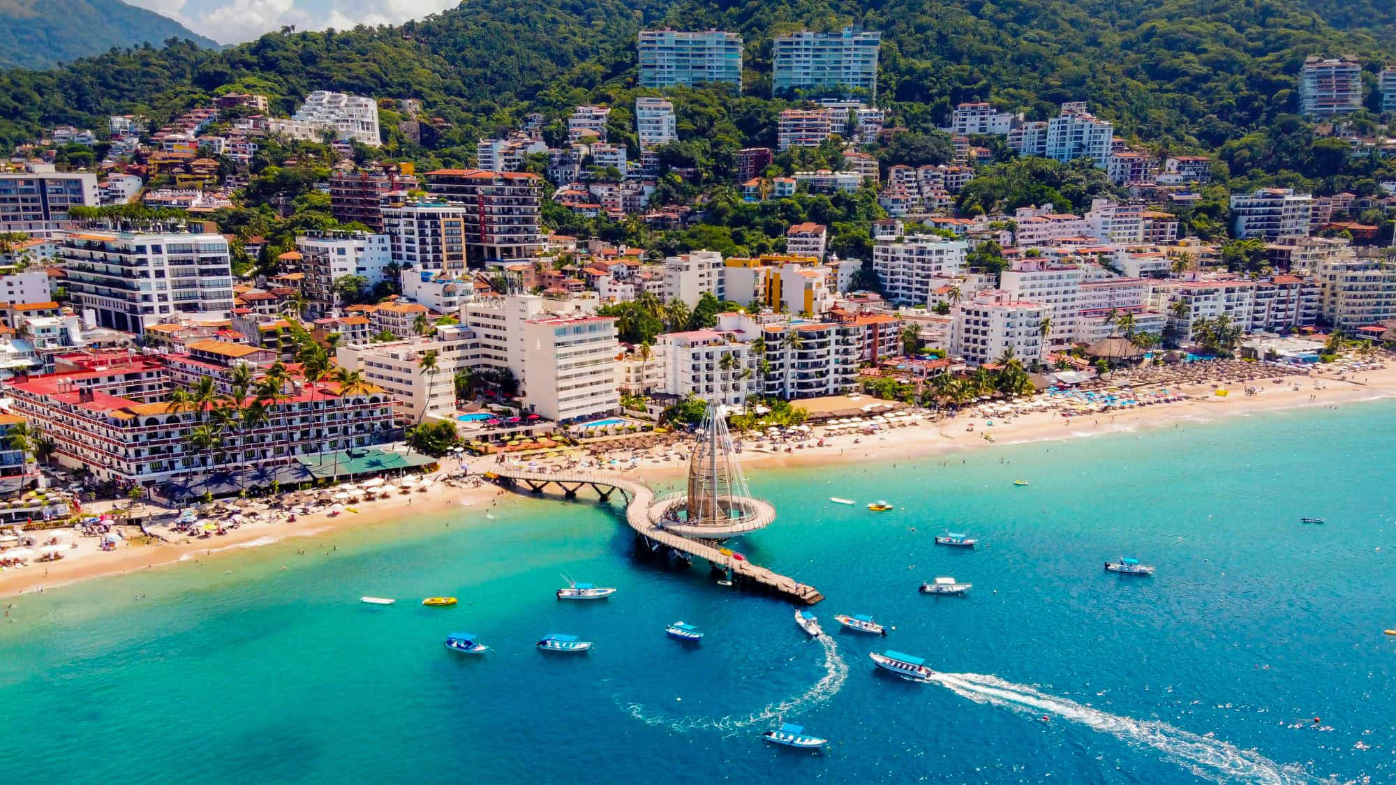 A scenic aerial shot of Puerto Vallarta's iconic pier extending into the blue-green ocean, surrounded by small boats with the lush hills and colorful buildings of the city in the background.