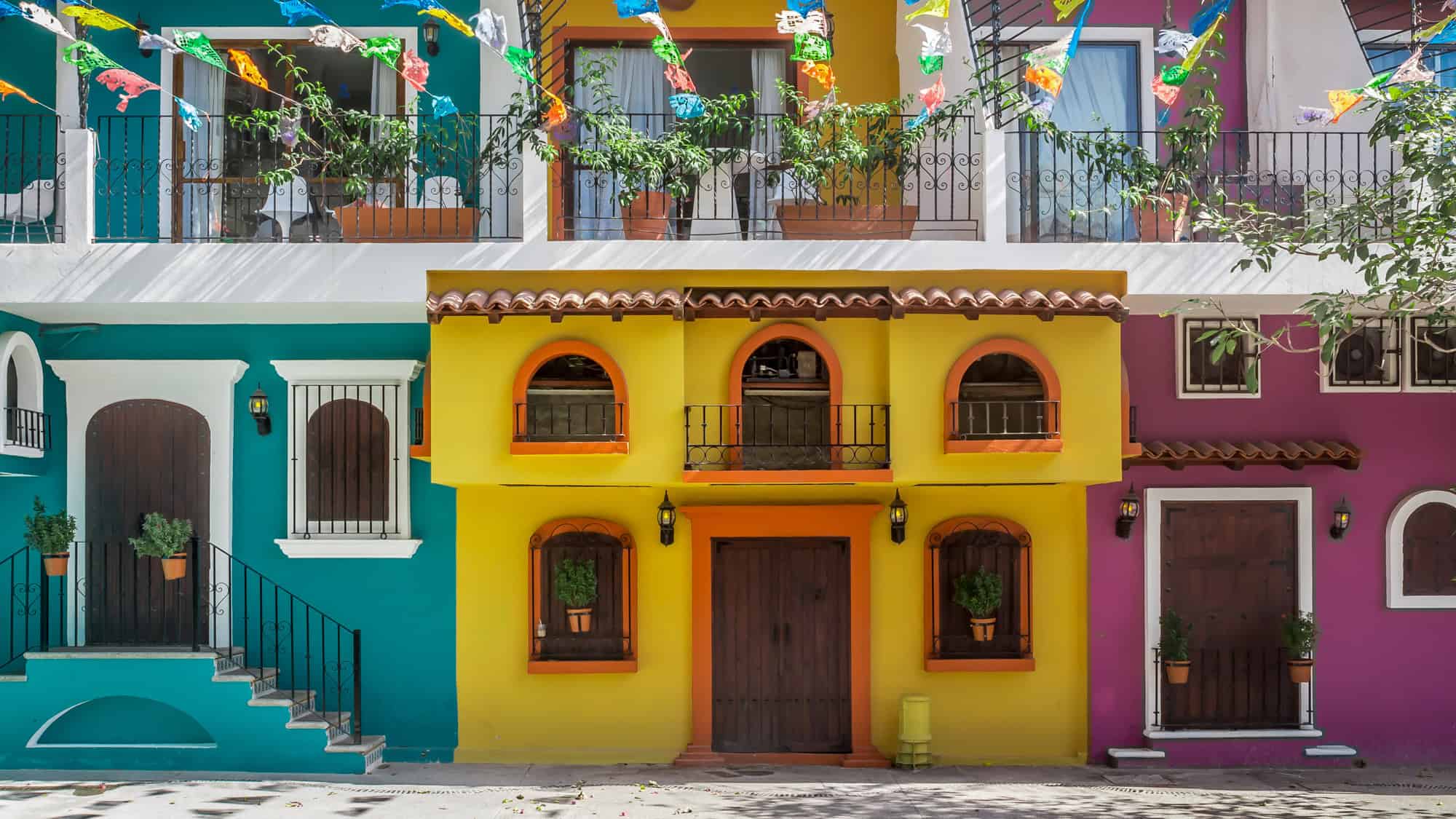 Brightly painted buildings in shades of teal, yellow, and magenta line a street, decorated with festive papel picado banners and potted plants in the windows.