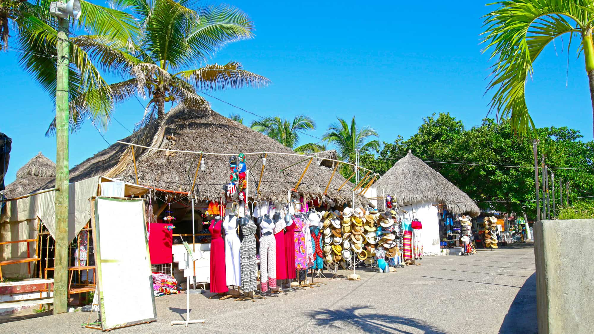 A colorful outdoor market in Puerto Vallarta, featuring vibrant dresses, hats, and souvenirs displayed under thatched-roof stalls with palm trees swaying overhead.