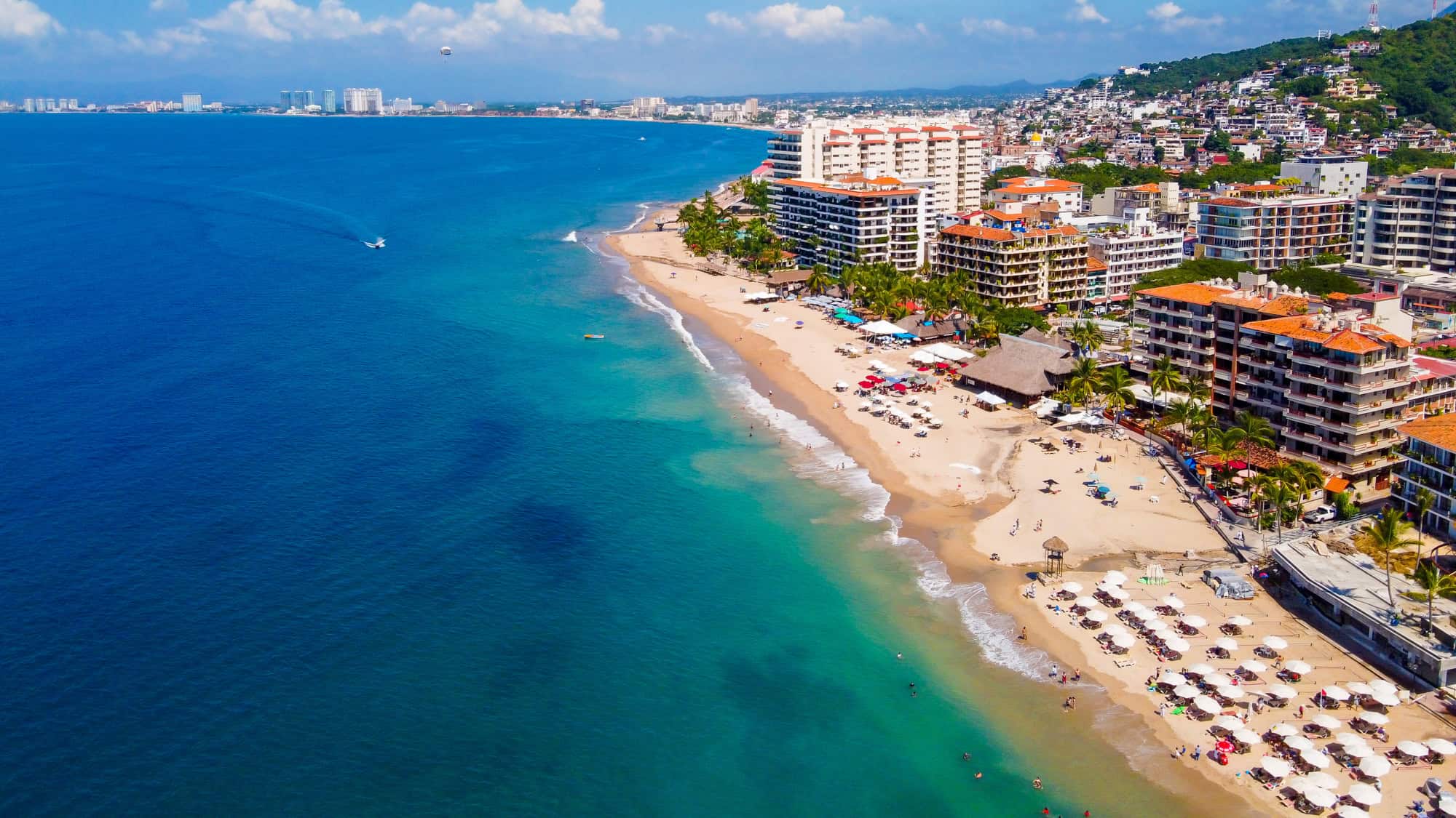 A vibrant aerial view of Puerto Vallarta's coastline, showcasing turquoise waters, sandy beaches lined with umbrellas, and tall hotels nestled between palm trees and city streets.