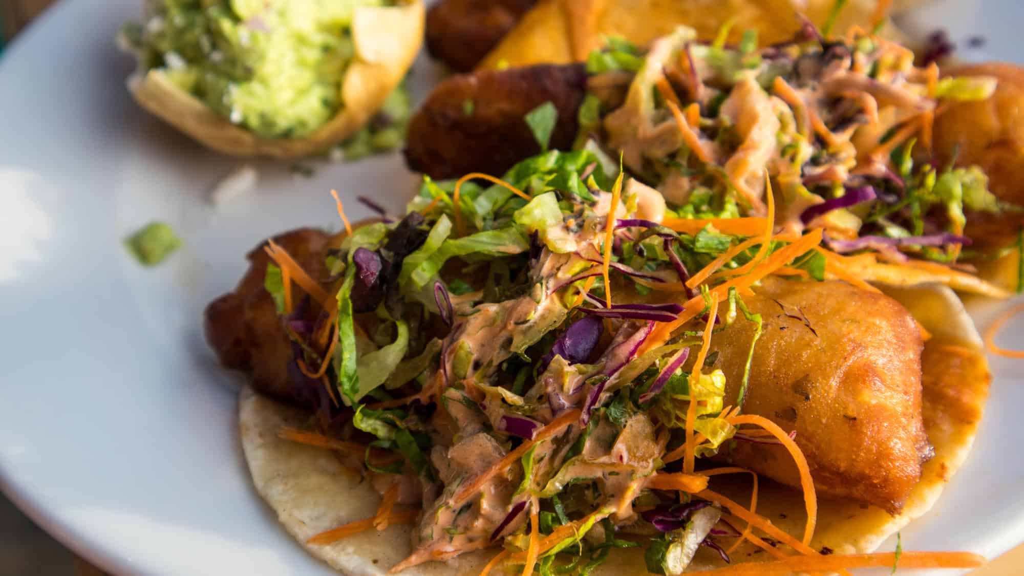 A close-up of crispy, golden fish tacos topped with shredded lettuce, carrots, and creamy sauce, served alongside guacamole and plantains on a white plate.
