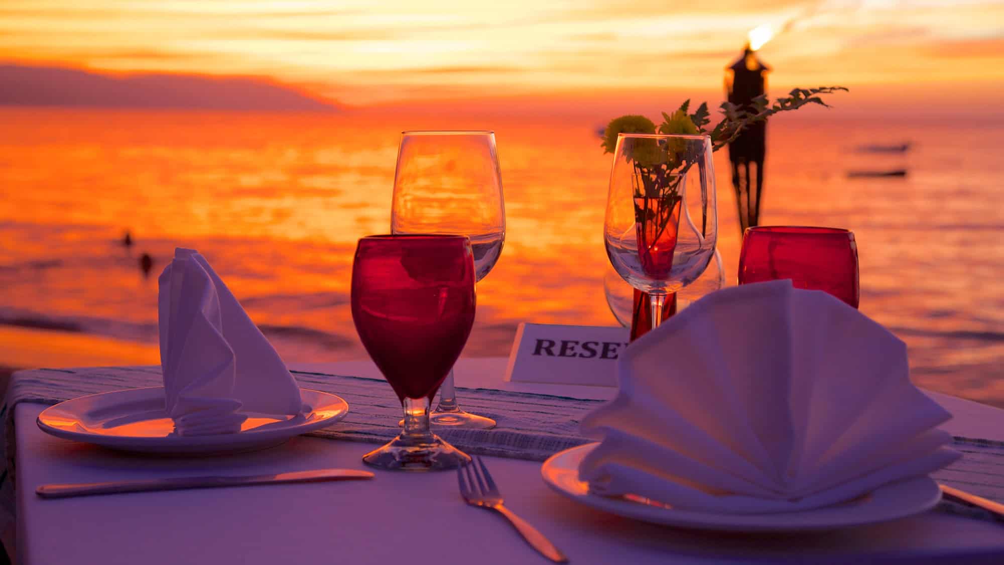 A romantic beachside dining table is set with folded napkins, glasses, and a "Reserved" sign, all glowing under a vibrant orange sunset.