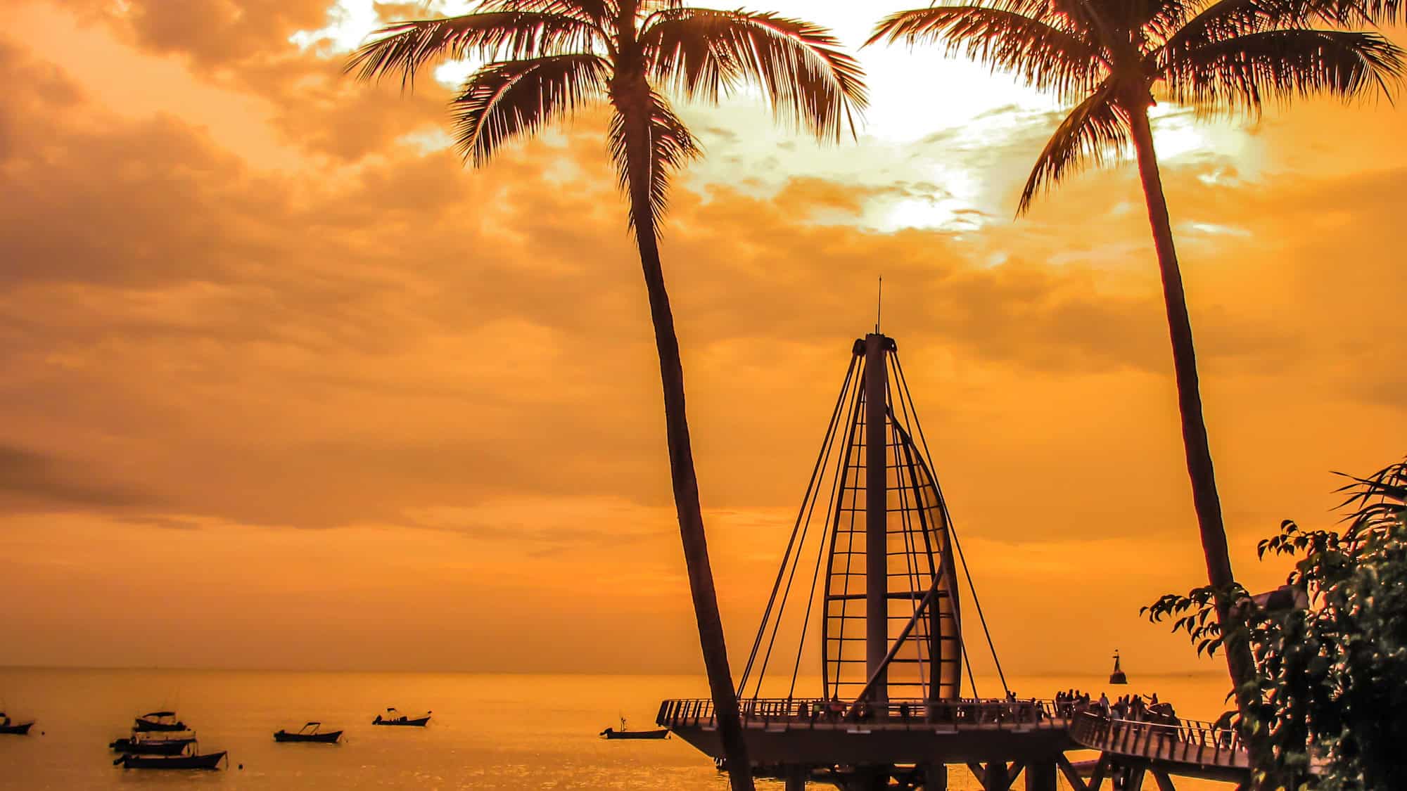 Silhouettes of palm trees and the Puerto Vallarta pier stand against a golden-orange sunset sky, with small boats floating peacefully on the calm water.