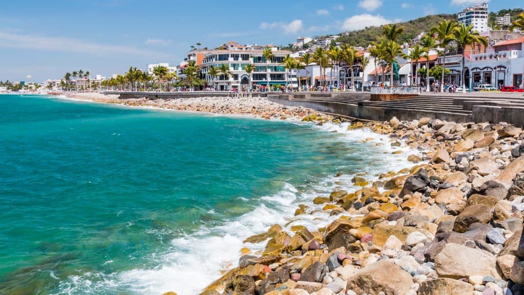 The Puerto Vallarta Malecón features a rocky shoreline with turquoise waves crashing against the coast, lined with palm trees and bustling waterfront buildings.