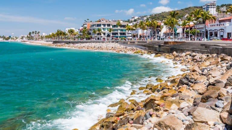The Puerto Vallarta Malecón features a rocky shoreline with turquoise waves crashing against the coast, lined with palm trees and bustling waterfront buildings.