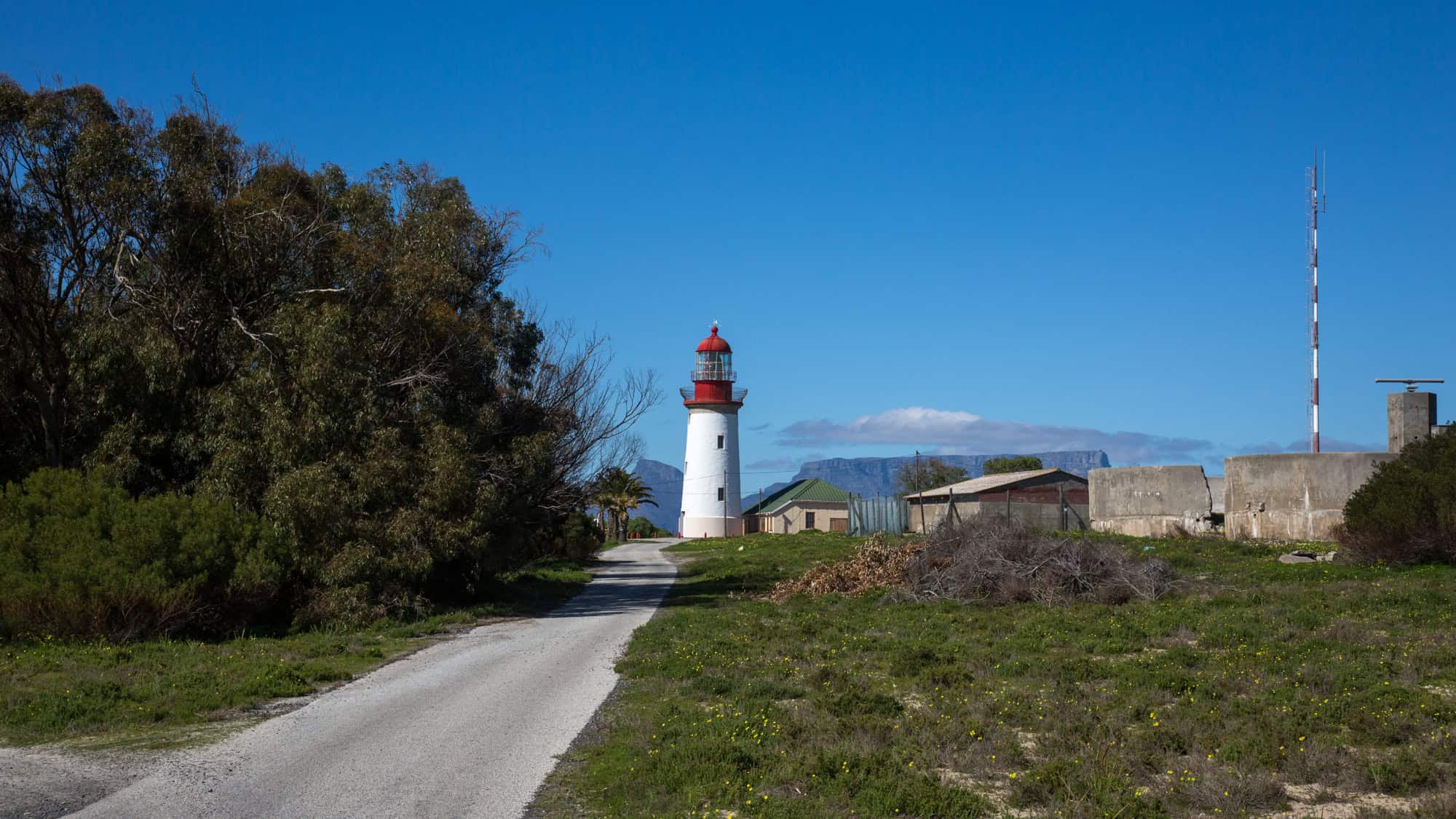 A serene coastal road leads to a white and red lighthouse, with Table Mountain faintly visible in the background. The rugged terrain and scattered buildings hint at the island’s historic significance.
