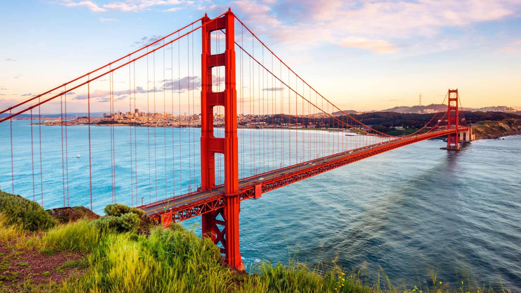 A scenic view of the Golden Gate Bridge in San Francisco, California, captured at sunset with the city skyline visible in the background and calm blue waters below.