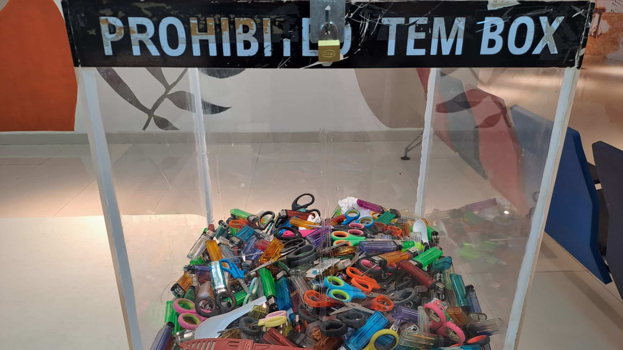 A transparent security checkpoint bin filled with confiscated items like scissors, lighters, and nail clippers. The top of the box has a sign that reads "PROHIBITED ITEM BOX."
