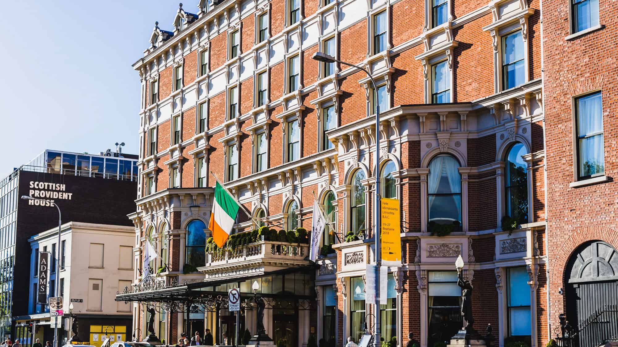 A grand red-brick hotel with ornate white window frames, featuring Irish flags waving at the entrance. The classic facade is lively, with city streets and pedestrians in the foreground.