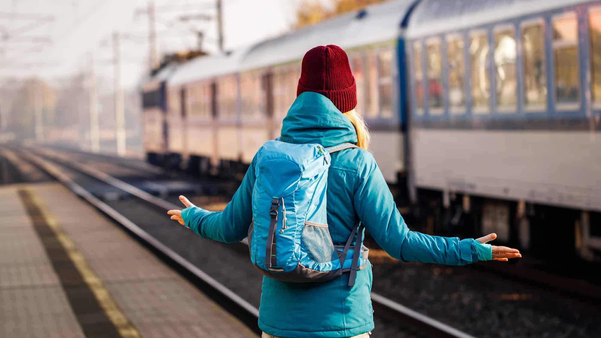A woman in a teal winter jacket and red beanie stands at an empty train platform, gesturing in confusion as a train departs in the background.