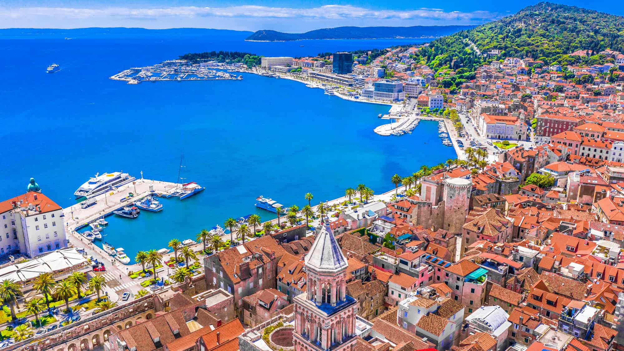 A stunning aerial view of Split, Croatia, featuring the city’s terracotta rooftops and the prominent bell tower of the Cathedral of Saint Domnius. The vibrant blue Adriatic Sea wraps around the harbor, dotted with yachts and cruise ships, while lush green hills frame the coastal cityscape.
