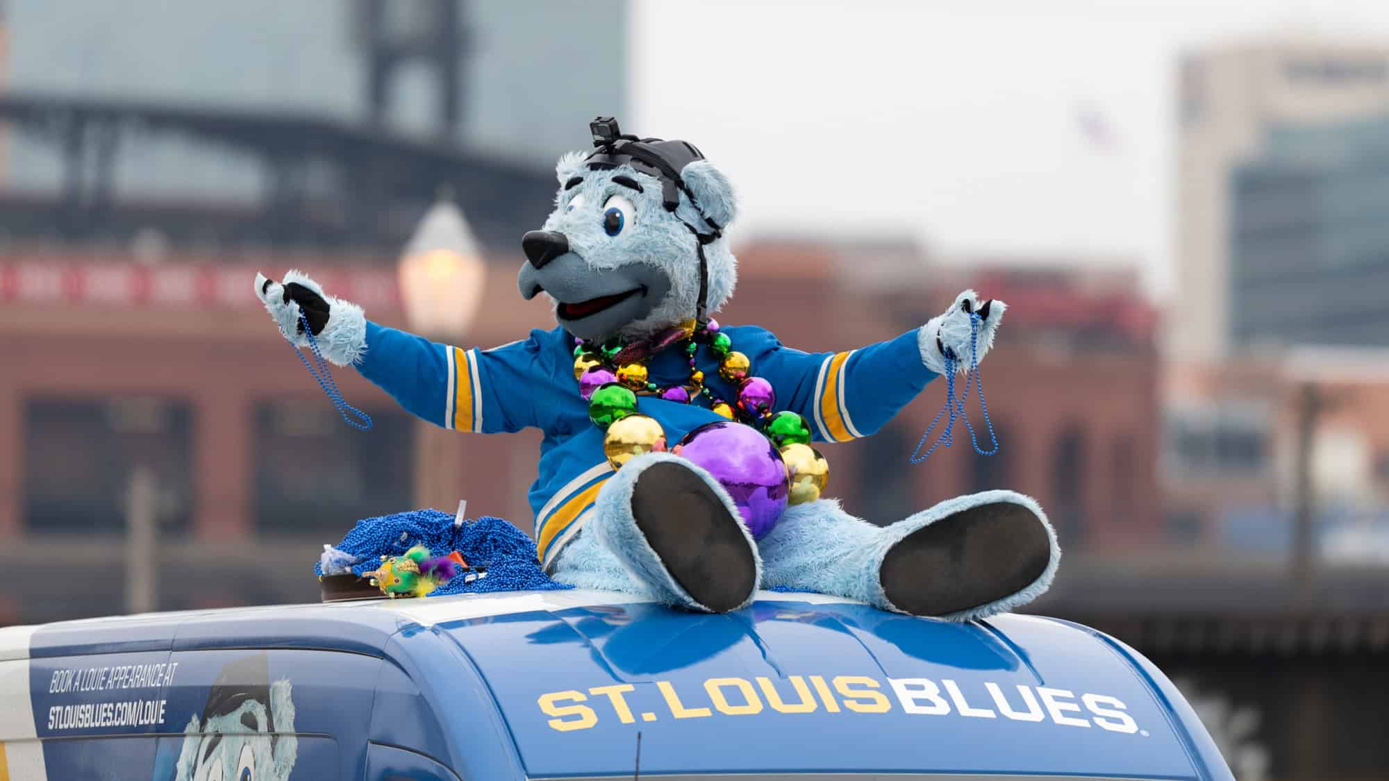 Louie, the St. Louis Blues mascot, sits atop a branded vehicle wearing Mardi Gras beads and holding blue strands, surrounded by festive decorations.