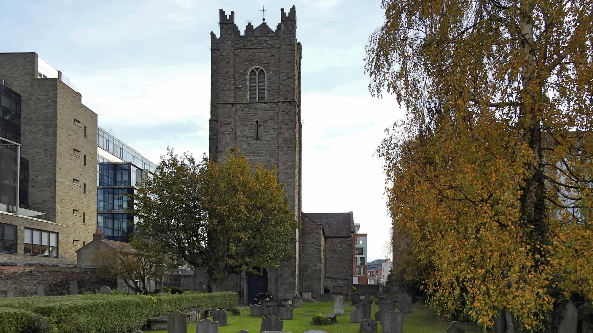 A medieval stone church with a tall tower rises above a graveyard filled with aged tombstones. Trees with golden autumn leaves frame the church, blending history with the surrounding urban landscape.