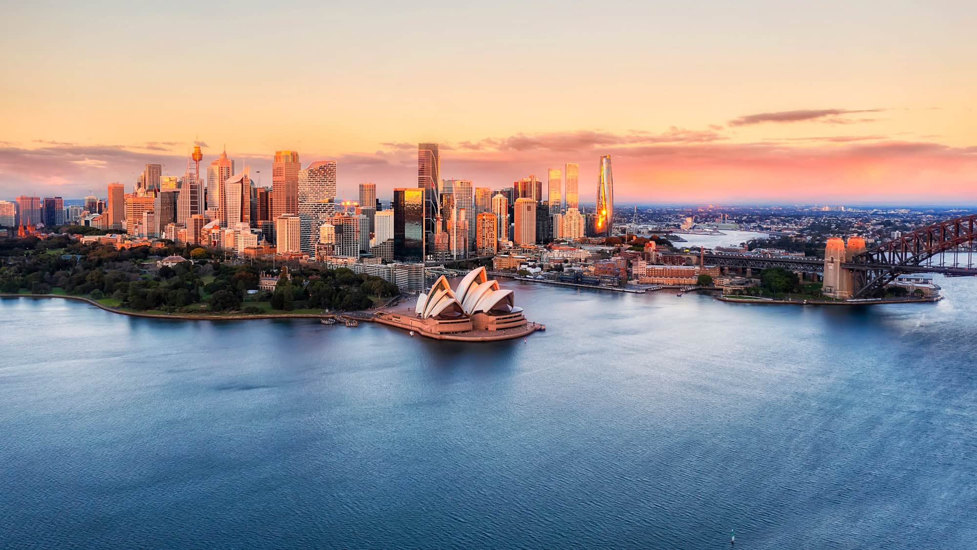 Aerial view of Sydney Harbour at sunset, featuring the iconic Sydney Opera House and Harbour Bridge with the city's skyline illuminated by the golden hour light.
