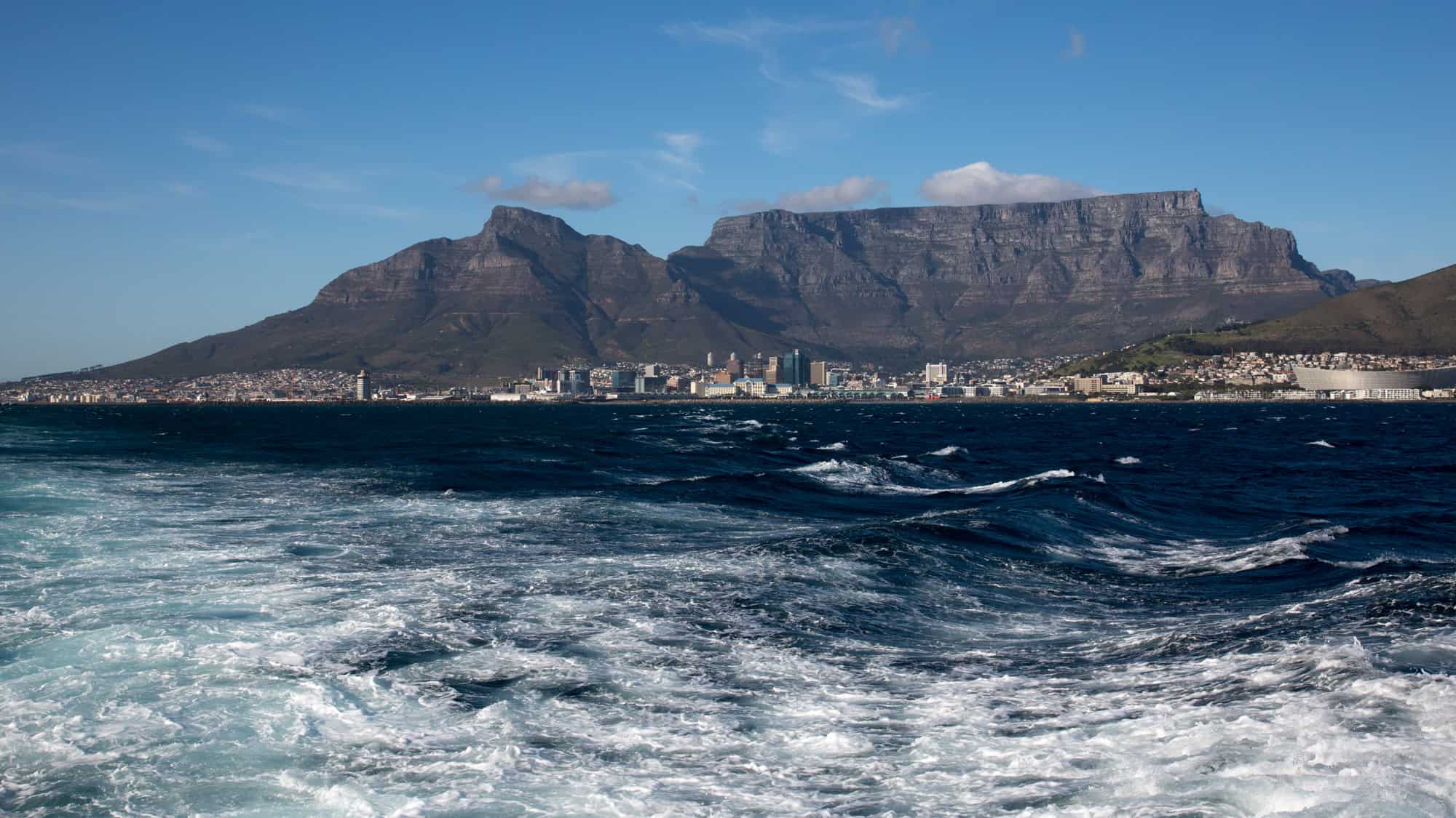 A breathtaking view of Cape Town with the iconic Table Mountain towering in the background. The deep blue ocean in the foreground has choppy waves, adding a dynamic contrast to the rugged mountain range.