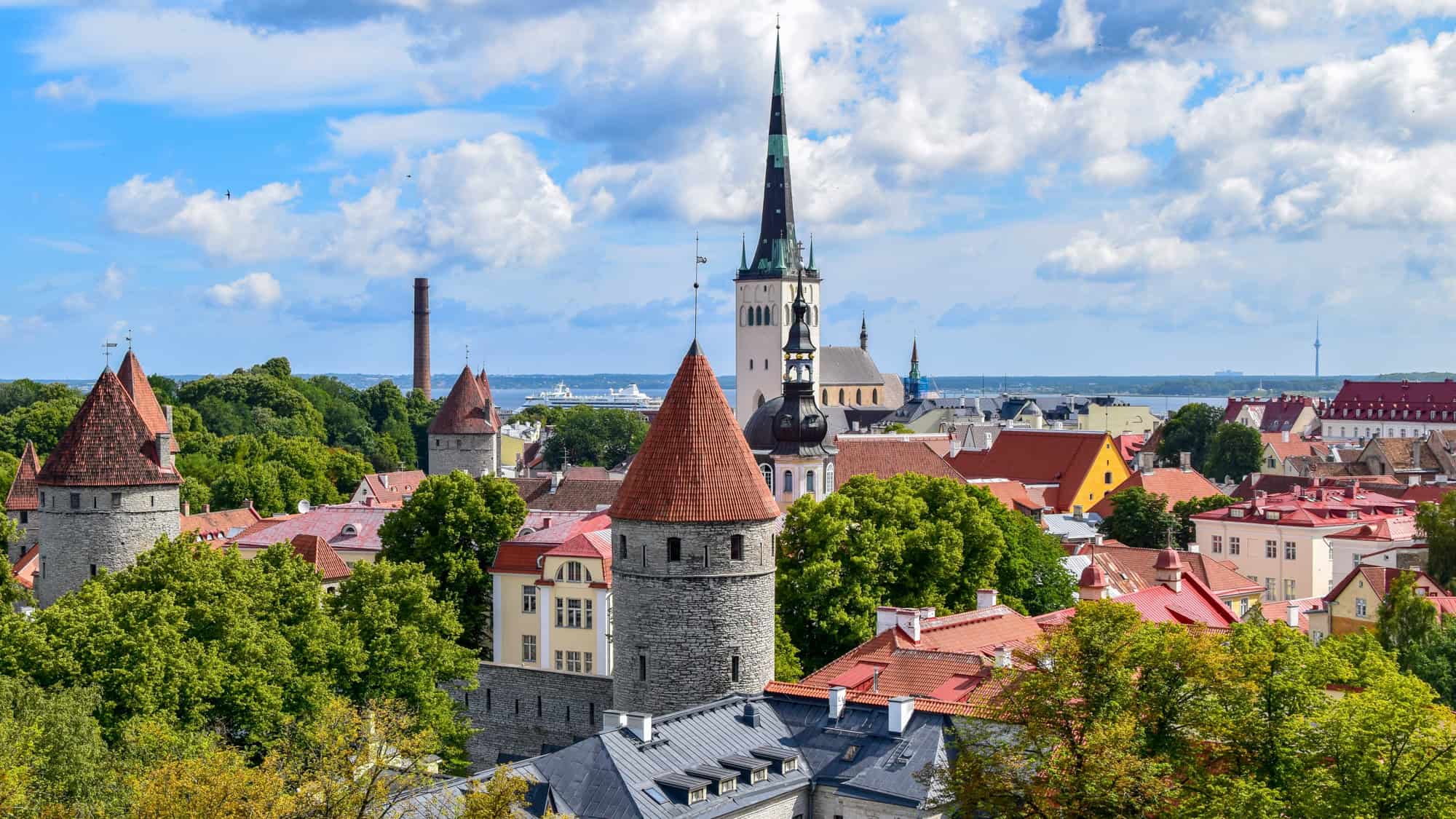 A picturesque view of Tallinn, Estonia, showcasing its medieval architecture with red-roofed stone towers and spires. St. Olaf’s Church rises prominently above the skyline, while leafy green trees add a fresh contrast to the historic city nestled by the Baltic Sea.