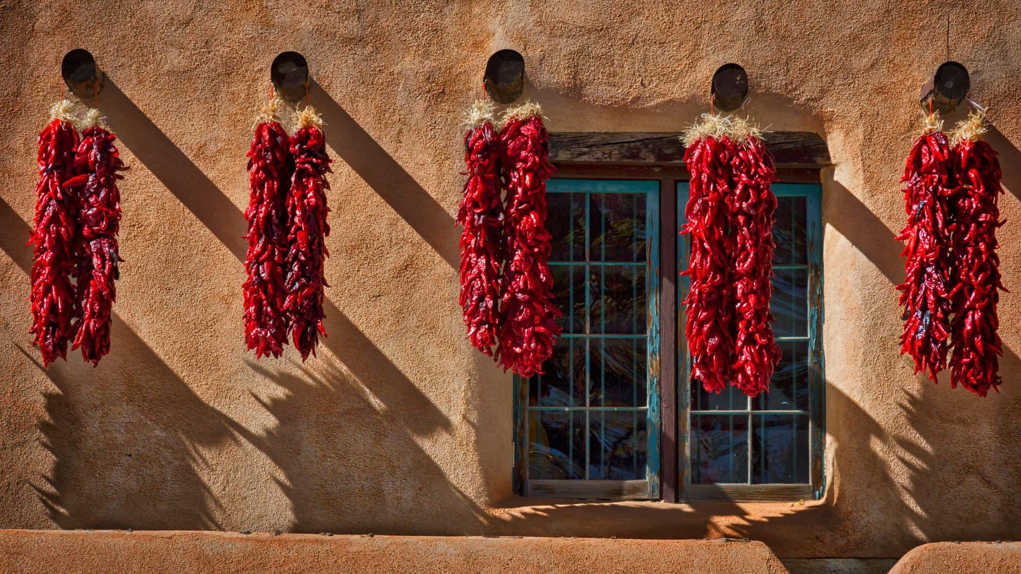 A rustic adobe wall in Taos, New Mexico, adorned with five vibrant red chili pepper ristras hanging to dry. The sun casts sharp shadows on the earthy-toned stucco, and a turquoise-framed window adds a pop of color.