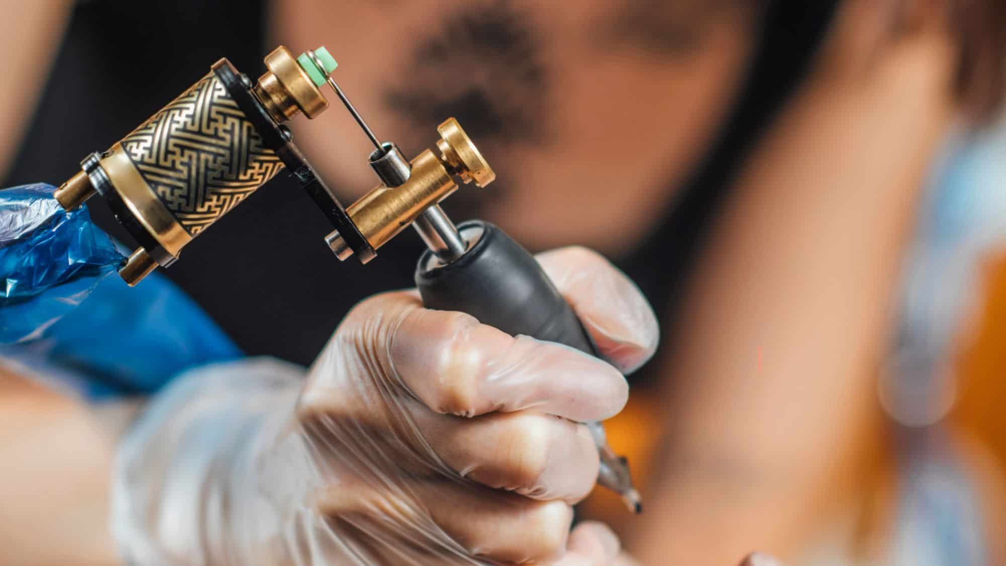 A close-up of a tattoo artist's gloved hands holding a tattoo machine with intricate golden detailing. The needle is in focus, ready for the next ink stroke.