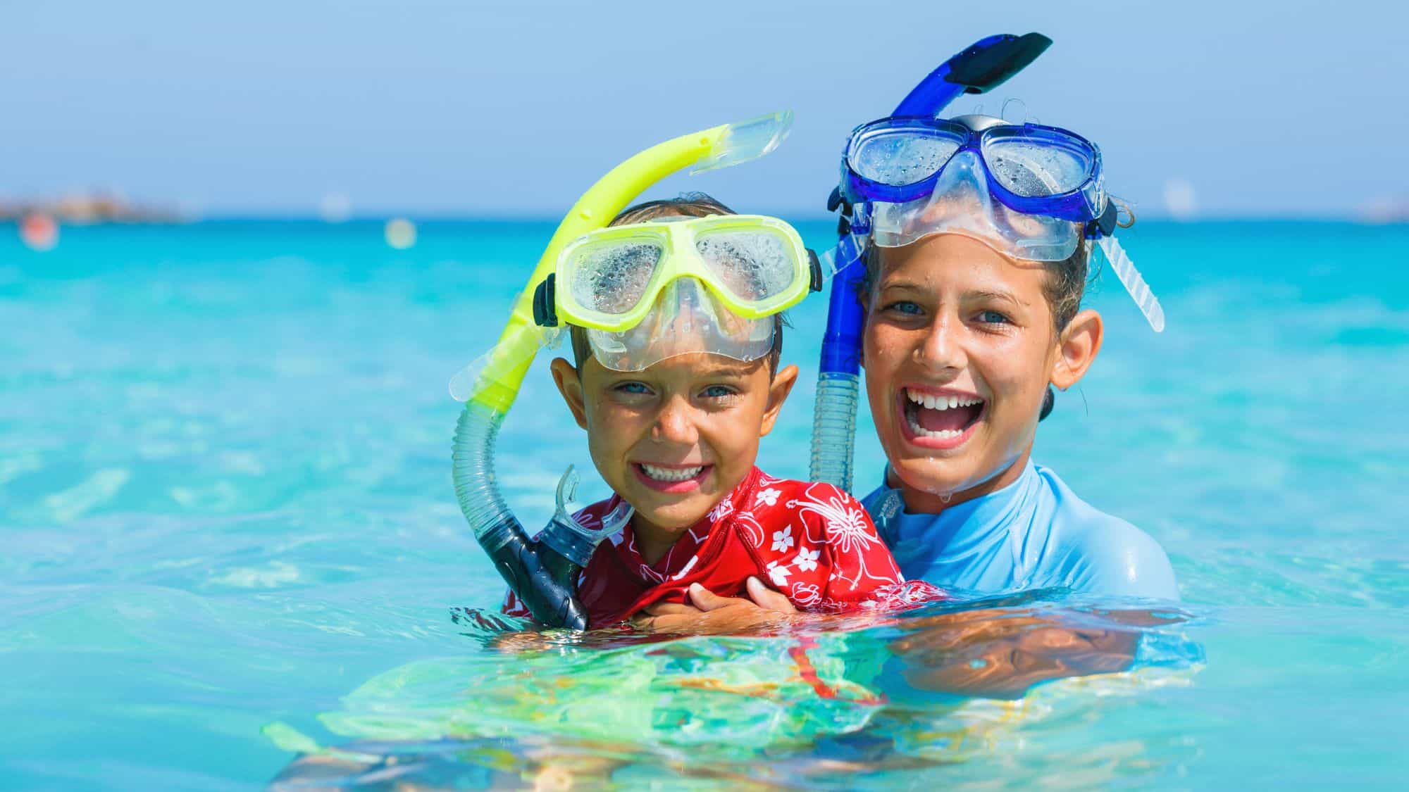 Two children in snorkeling gear, one in a red rash guard and the other in a blue one, are grinning while partially submerged in crystal-clear turquoise water. Their goggles are wet, and they appear to be enjoying a snorkeling adventure.