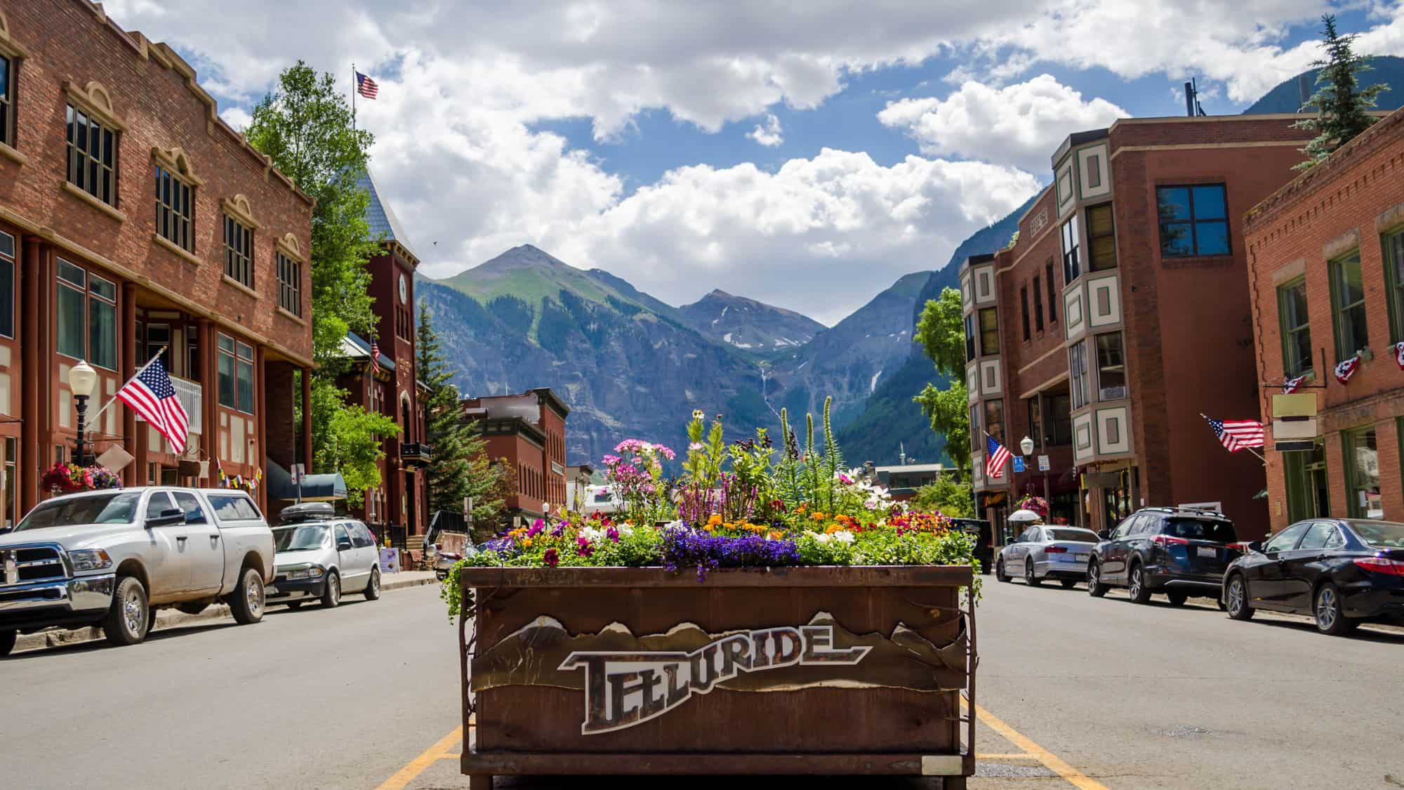 A picturesque street view of Telluride, Colorado, with a planter filled with colorful flowers labeled "Telluride" in the foreground. Historic brick buildings line the street, with towering, snow-capped mountains rising in the background.