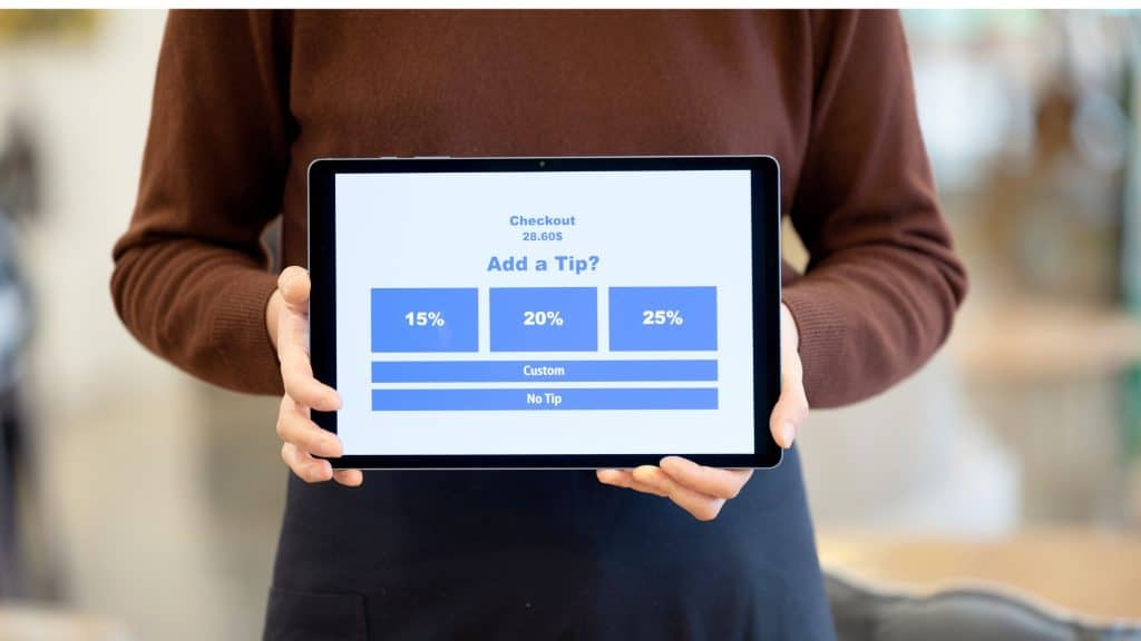A person wearing a brown sweater holds up a digital tablet displaying a tipping screen with options for 15%, 20%, 25%, a custom amount, or no tip at all. The screen shows a checkout total of 28.60.