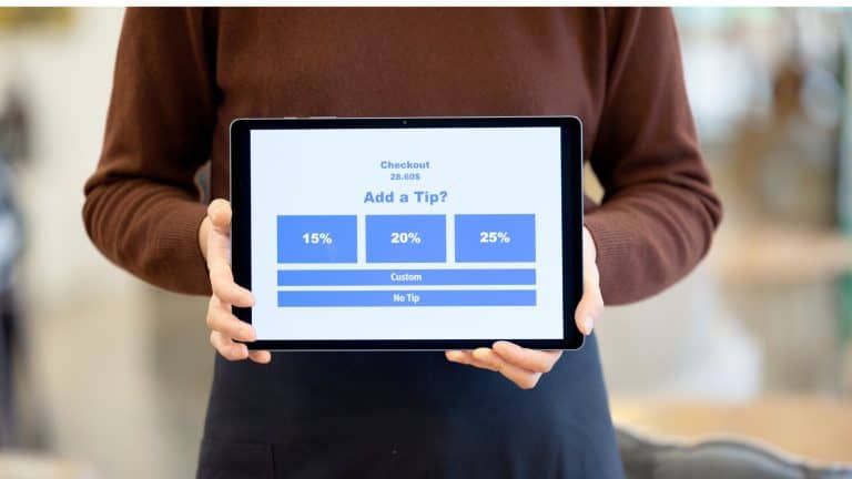A person wearing a brown sweater holds up a digital tablet displaying a tipping screen with options for 15%, 20%, 25%, a custom amount, or no tip at all. The screen shows a checkout total of 28.60.