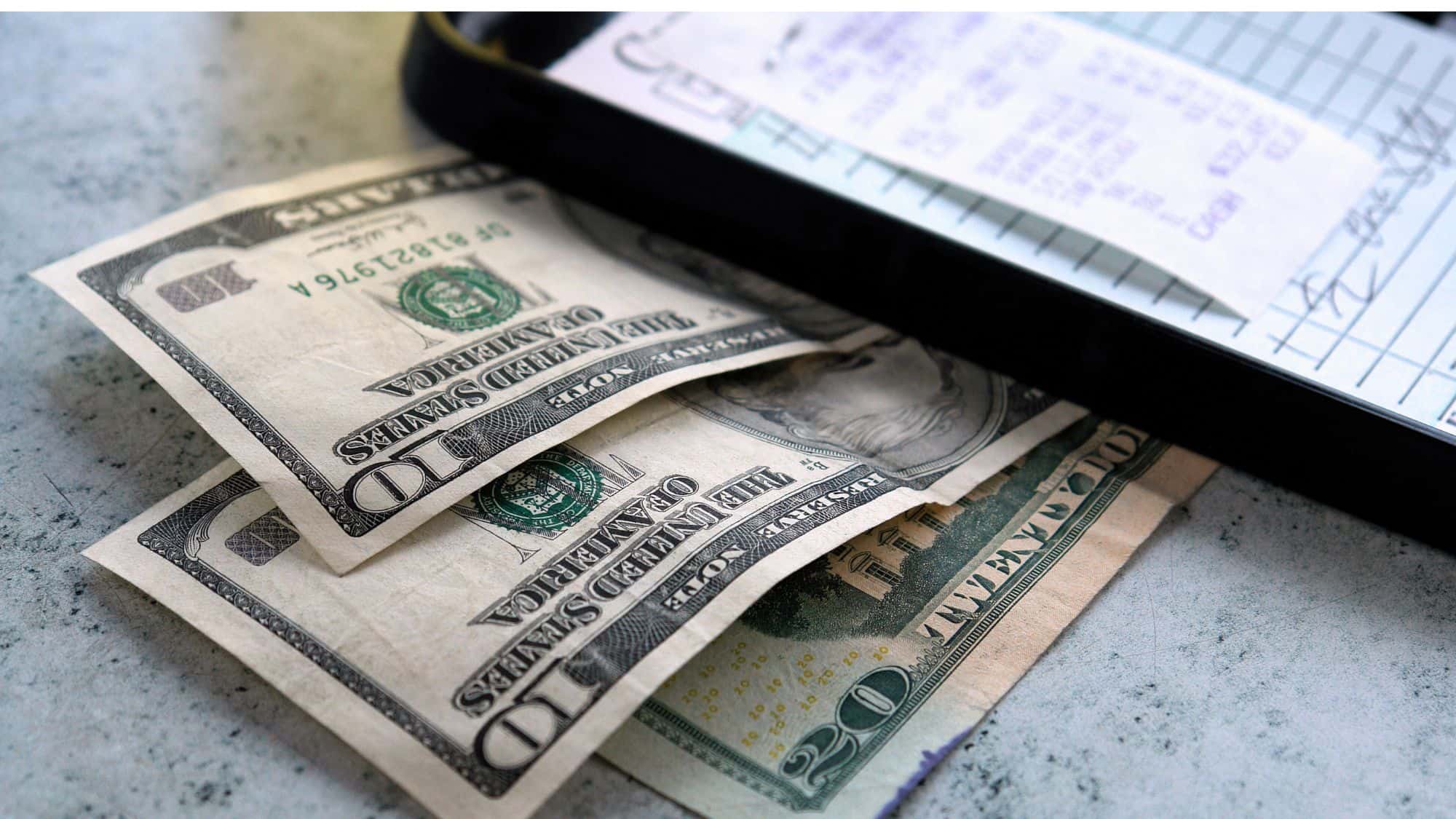 A close-up of a restaurant bill on a black check presenter with several US dollar bills placed on top, suggesting a cash tip.